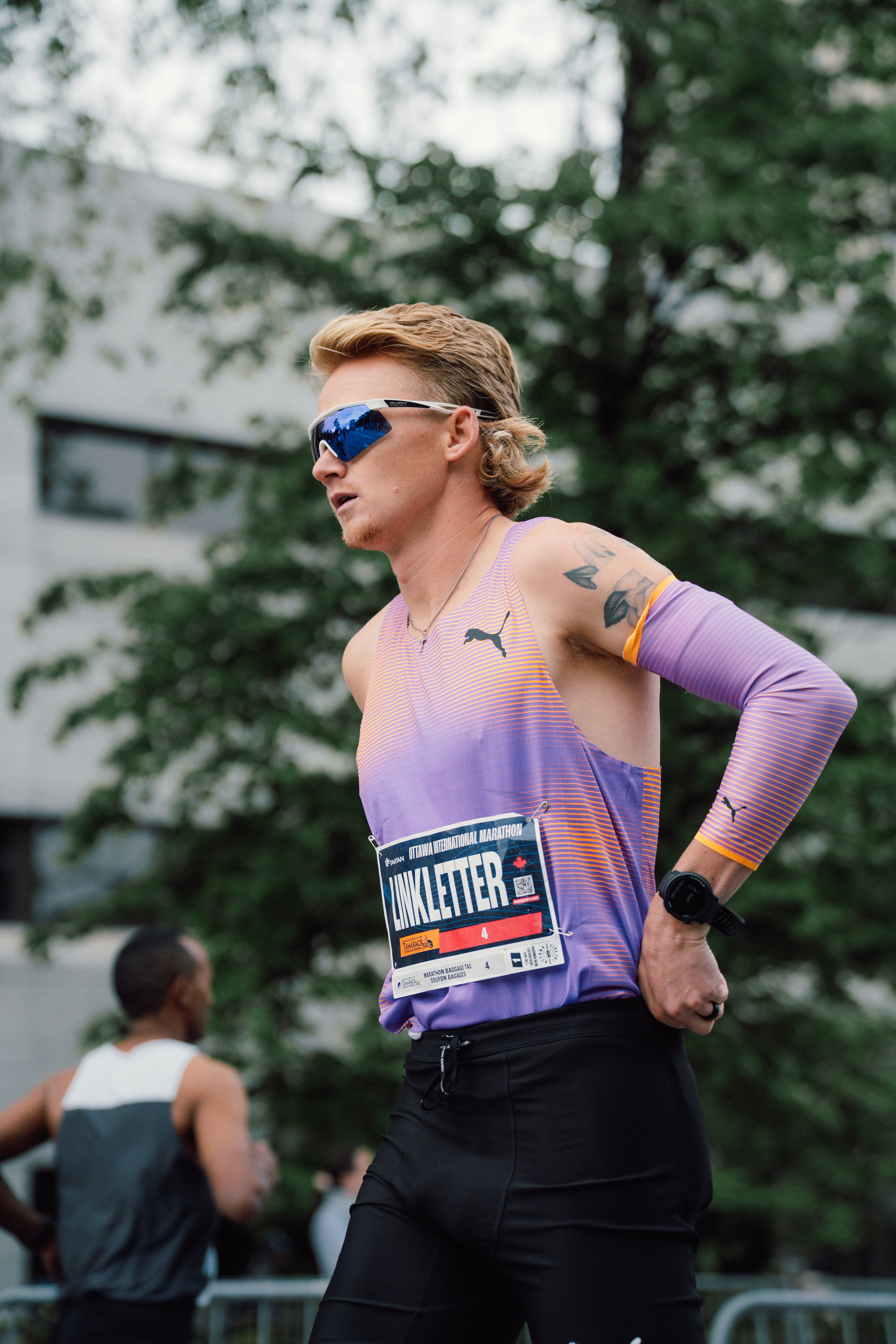 Rory Linkletter wearing sunglasses, a purple and orange athletic shirt, and a bib number, standing with hands on hips during a race.