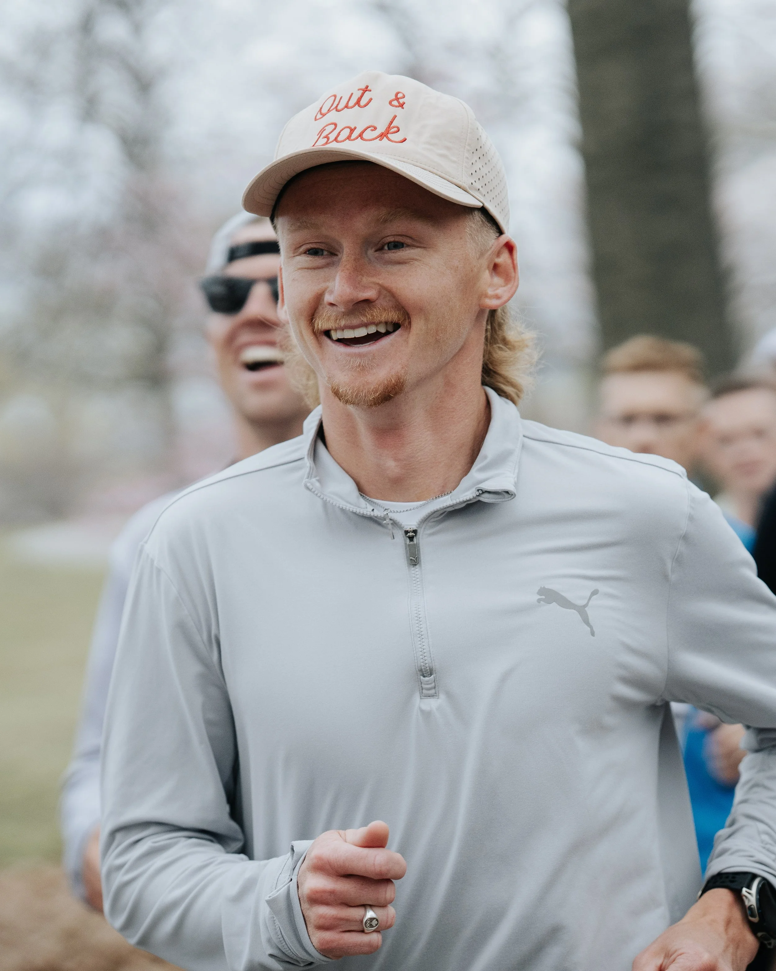 A close-up of Rory Linkletter running outdoors in a park or wooded area, wearing a beige cap with red script reading 'out & Back' and a light gray zip-up athletic jacket with a Puma logo.