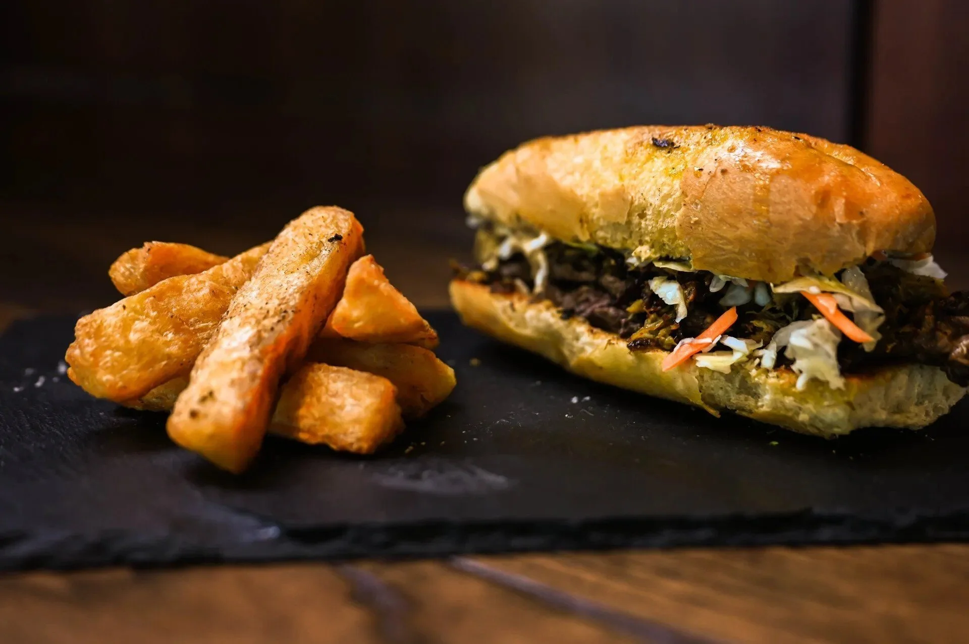 A sandwich with shredded vegetables and meat on a bun, served with potato wedges on a black slate plate.