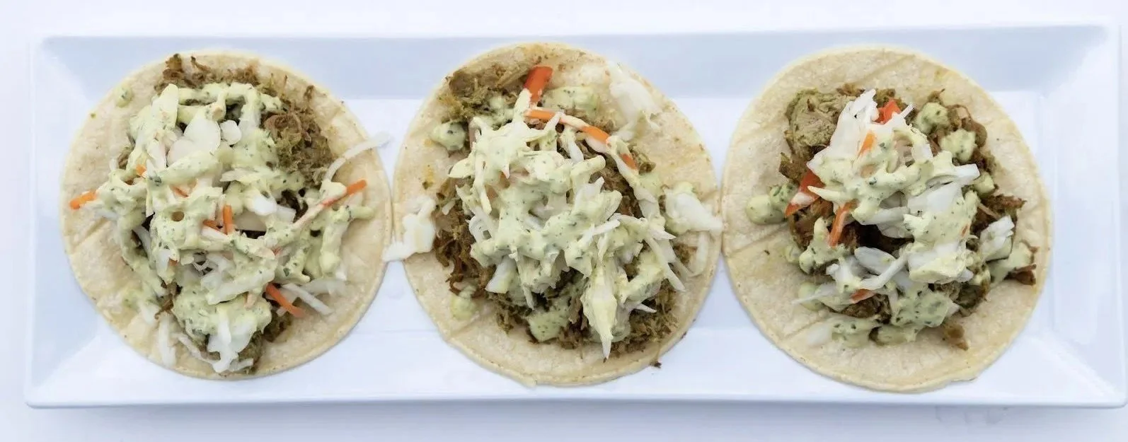 Three tacos with shredded meat, cabbage, and sauce on a white rectangular platter.
