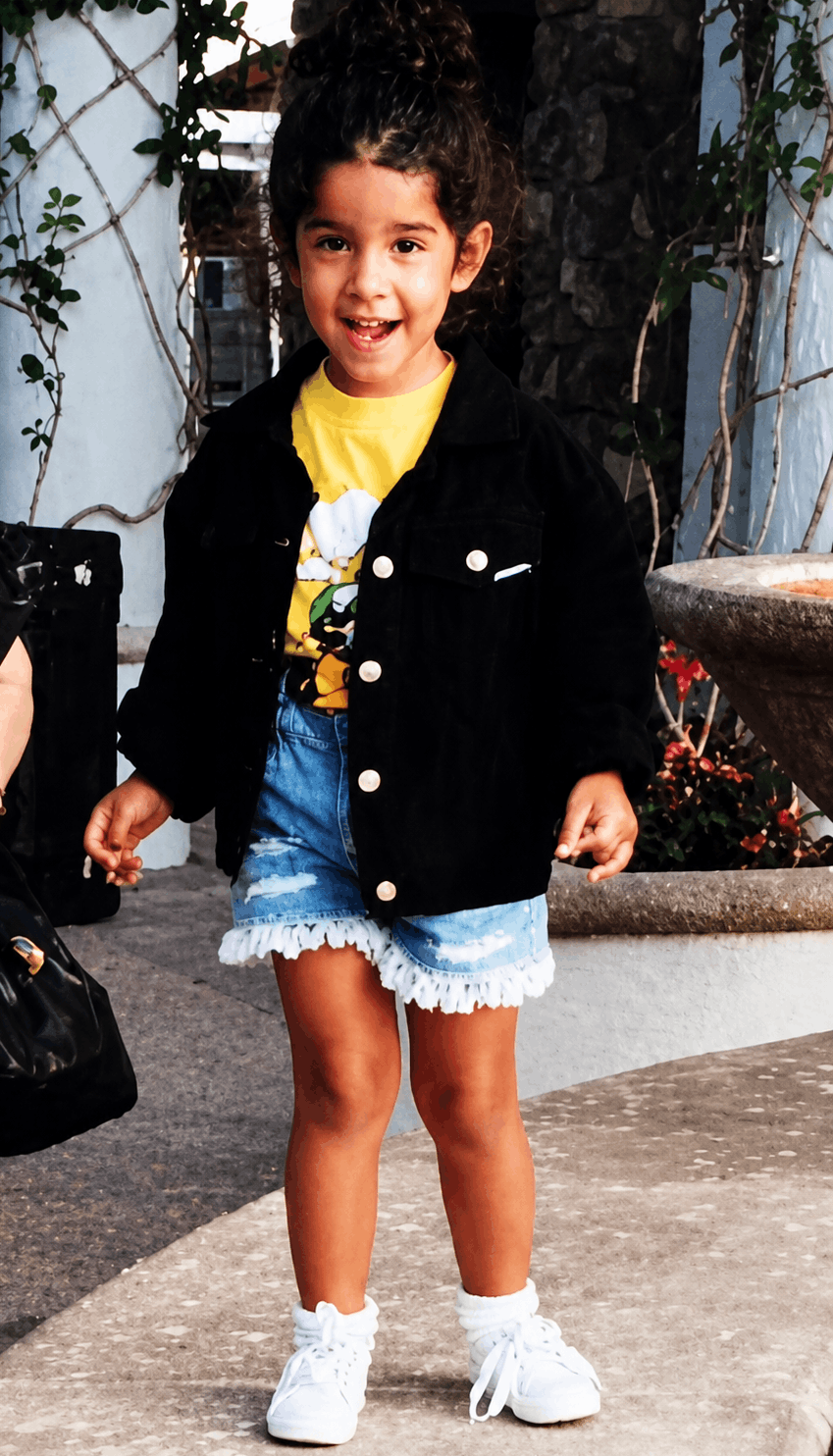 A young Nadia with curly hair smiling, wearing a yellow T-shirt, a black jacket, denim shorts with ruffled edges, white sneakers, and white socks, standing outdoors near a stone fountain and surrounded by plants.