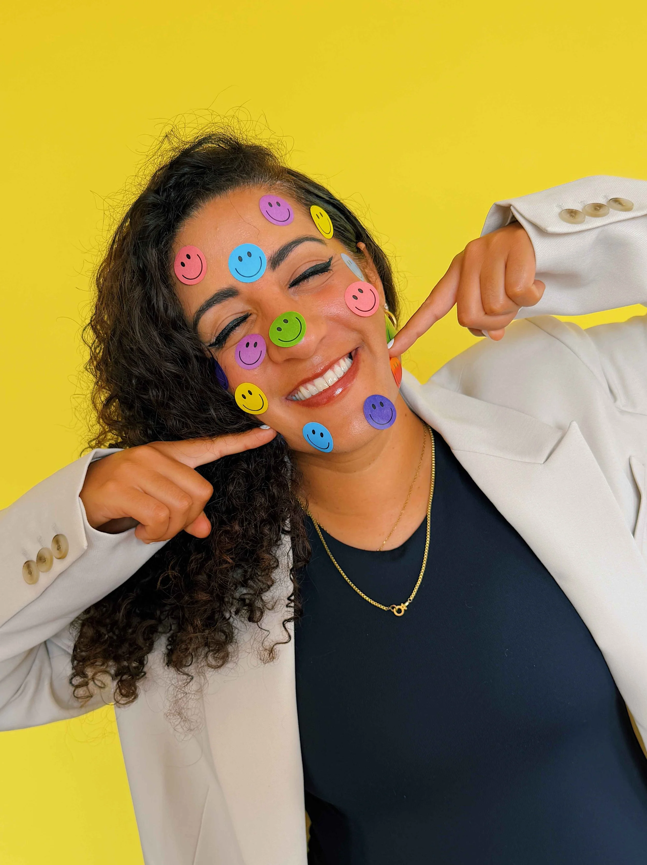 Nadia Zadeh (founder of MND MTTRS) with curly hair smiling and joyfully touching her face, covered with colorful smiley face stickers, against a bright yellow background, dressed in a white blazer and a dark top.