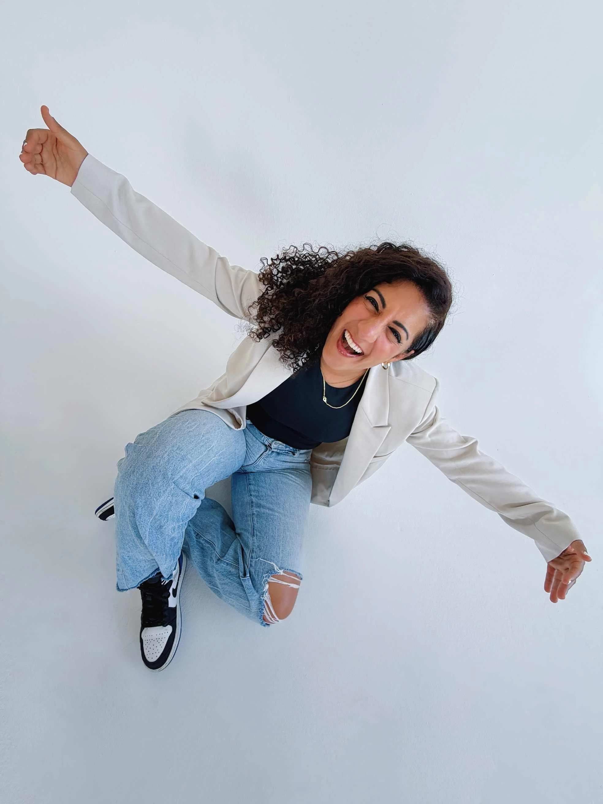 Woman with curly hair smiling and posing playfully, wearing a beige blazer, black top, ripped jeans, and sneakers, against a plain white background.