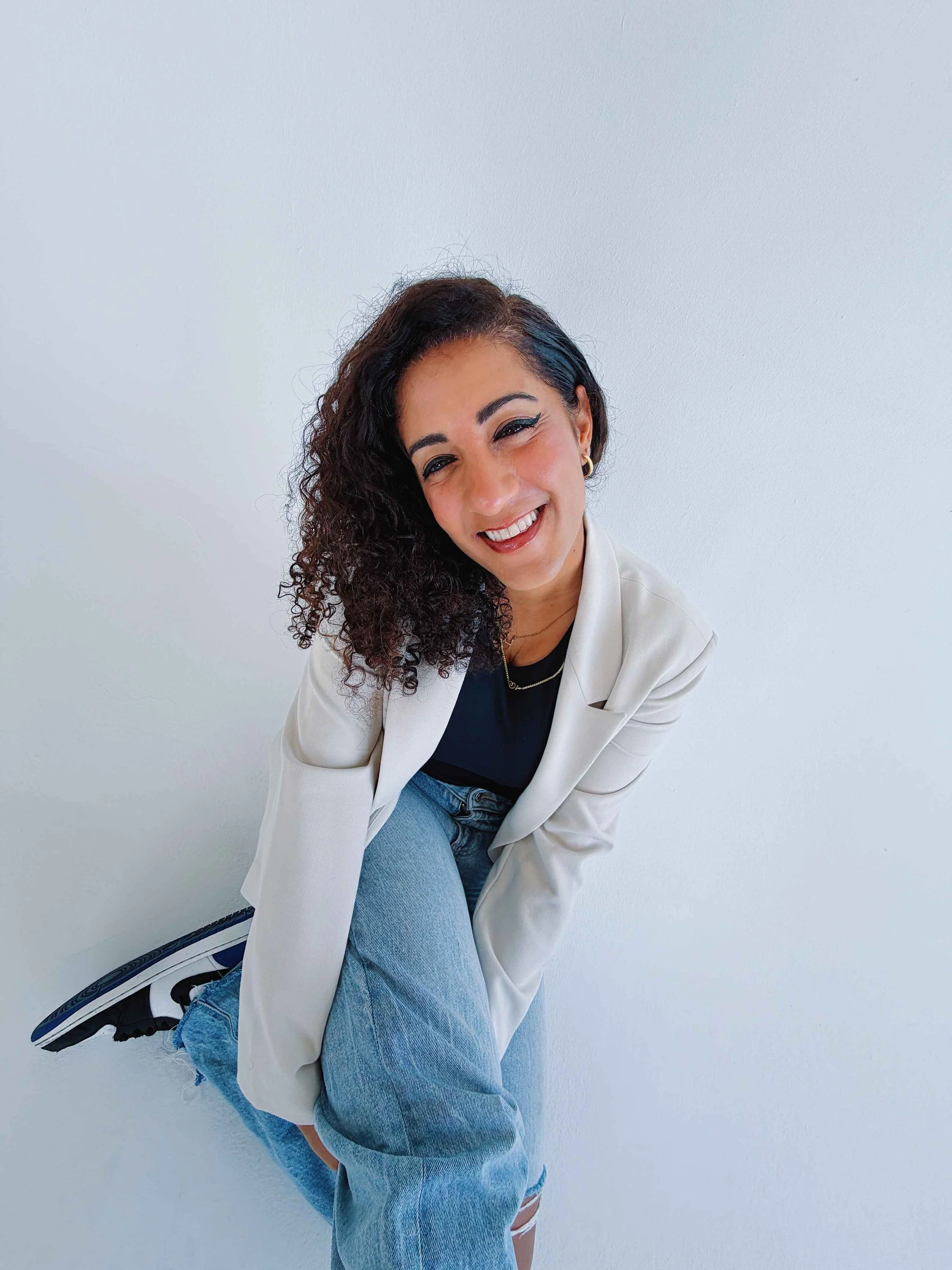 A woman with curly dark hair and earrings, wearing a white blazer, black top, and light blue ripped jeans, smiling and squatting against a plain light-colored wall.