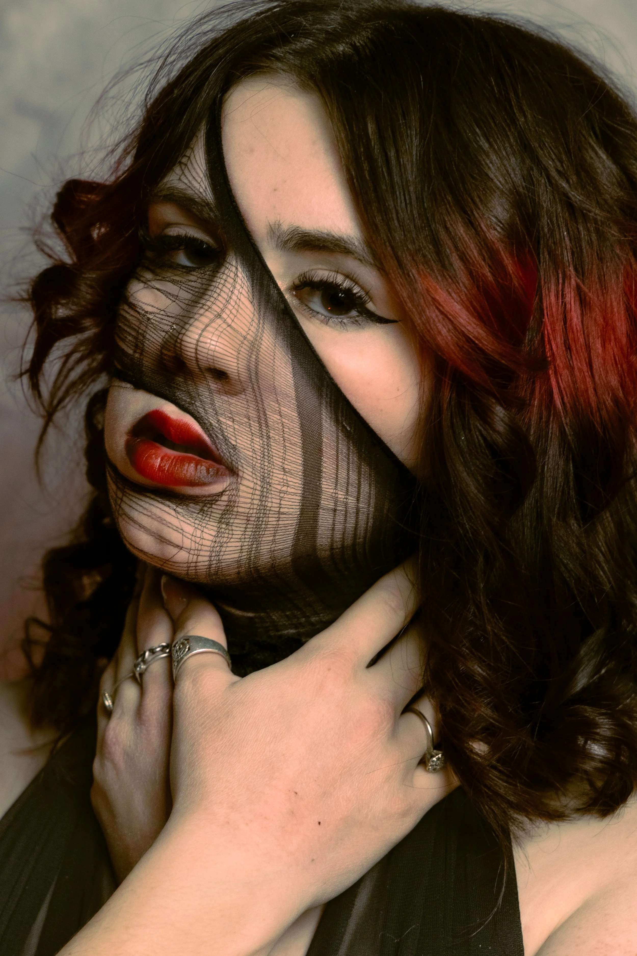 A woman with half of her face obscured by a black, lace fabric with a paisley pattern, revealing her eye, eyebrow, and makeup. She has long, wavy brown hair with red highlights, dark eyeliner, and red lipstick. Both her hands are visible, with rings on her fingers.