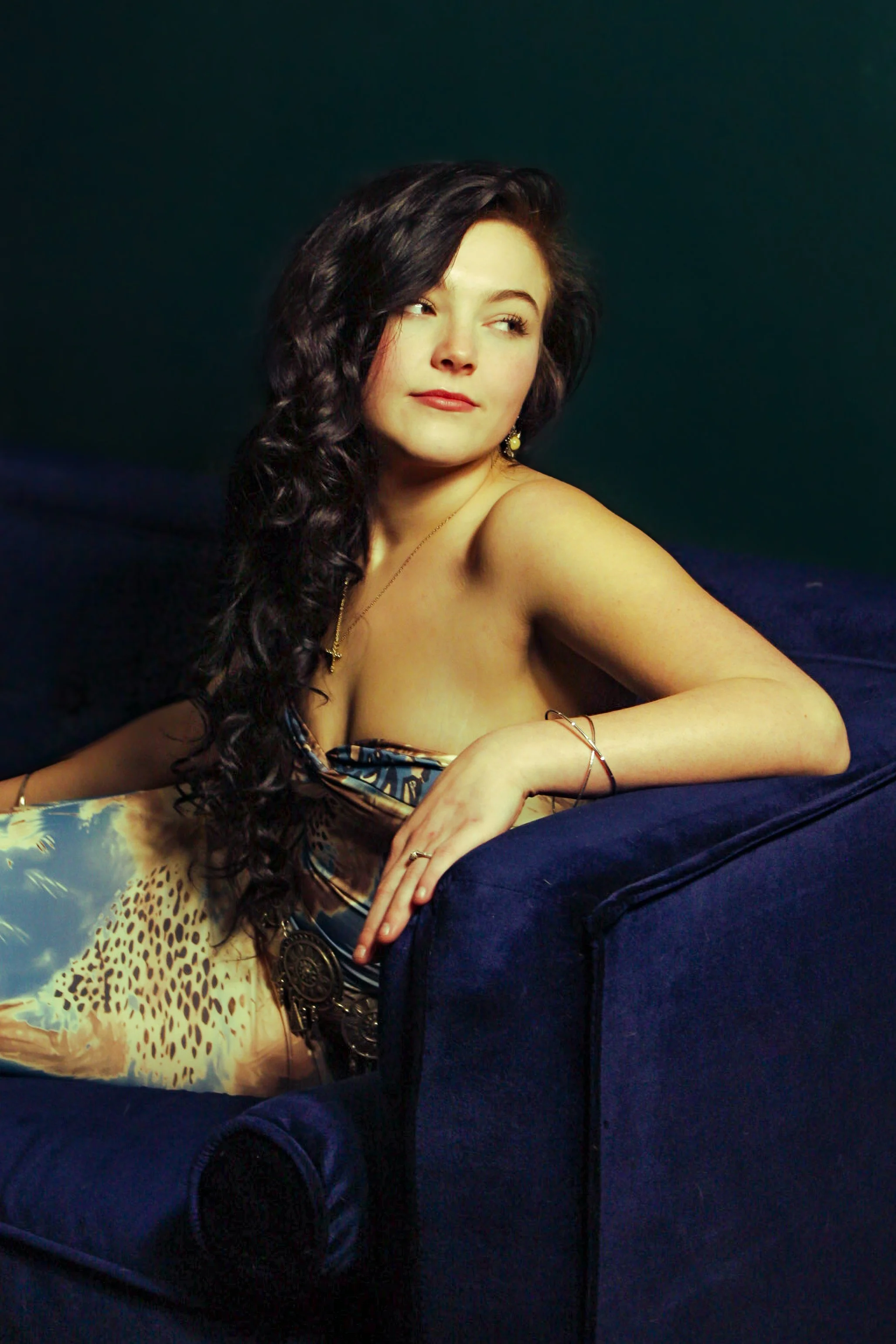 A woman with long dark curly hair, wearing a dress with a leopard print pattern, is sitting on a dark blue couch.