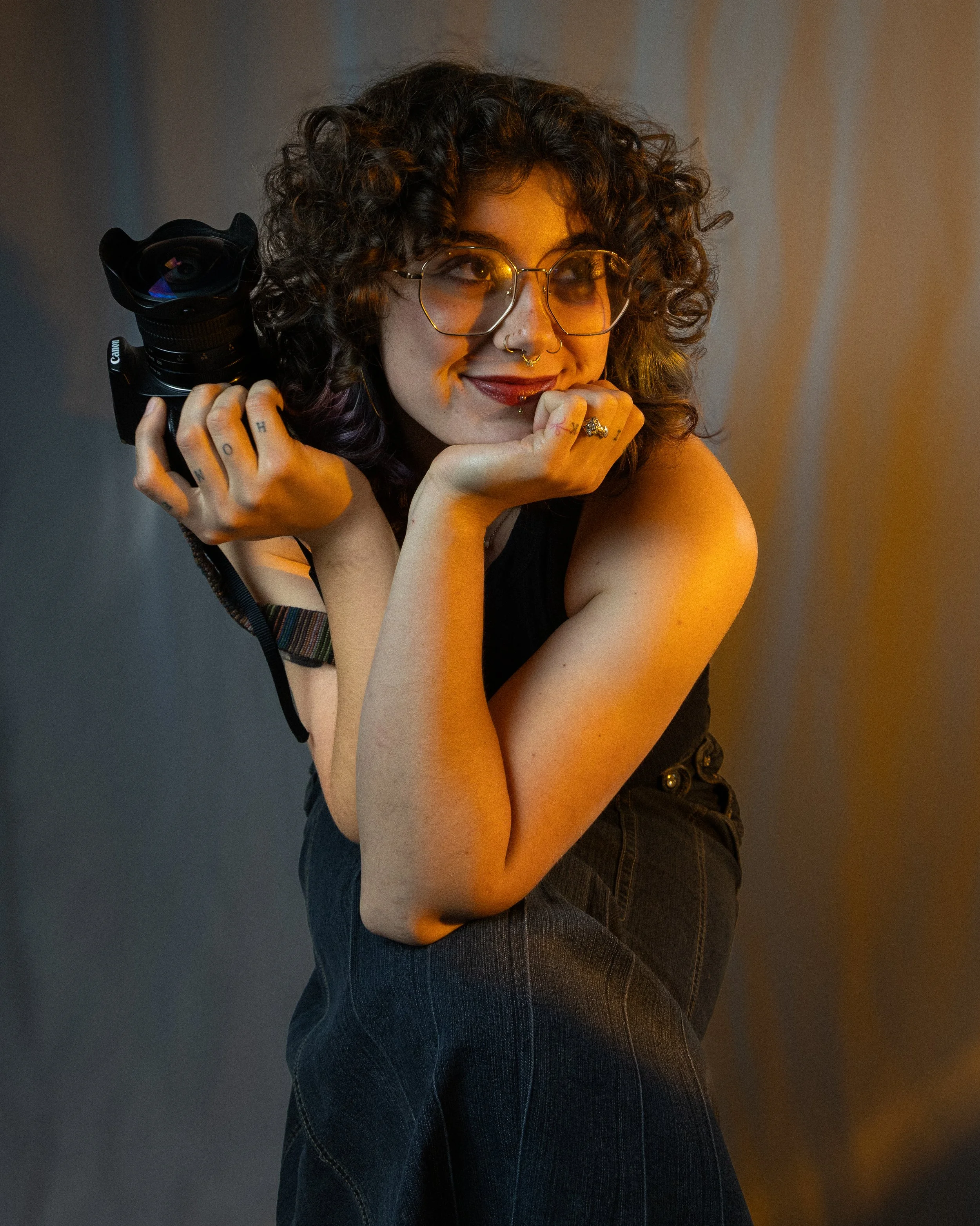 A woman with curly hair and glasses holding a camera against a dark background.