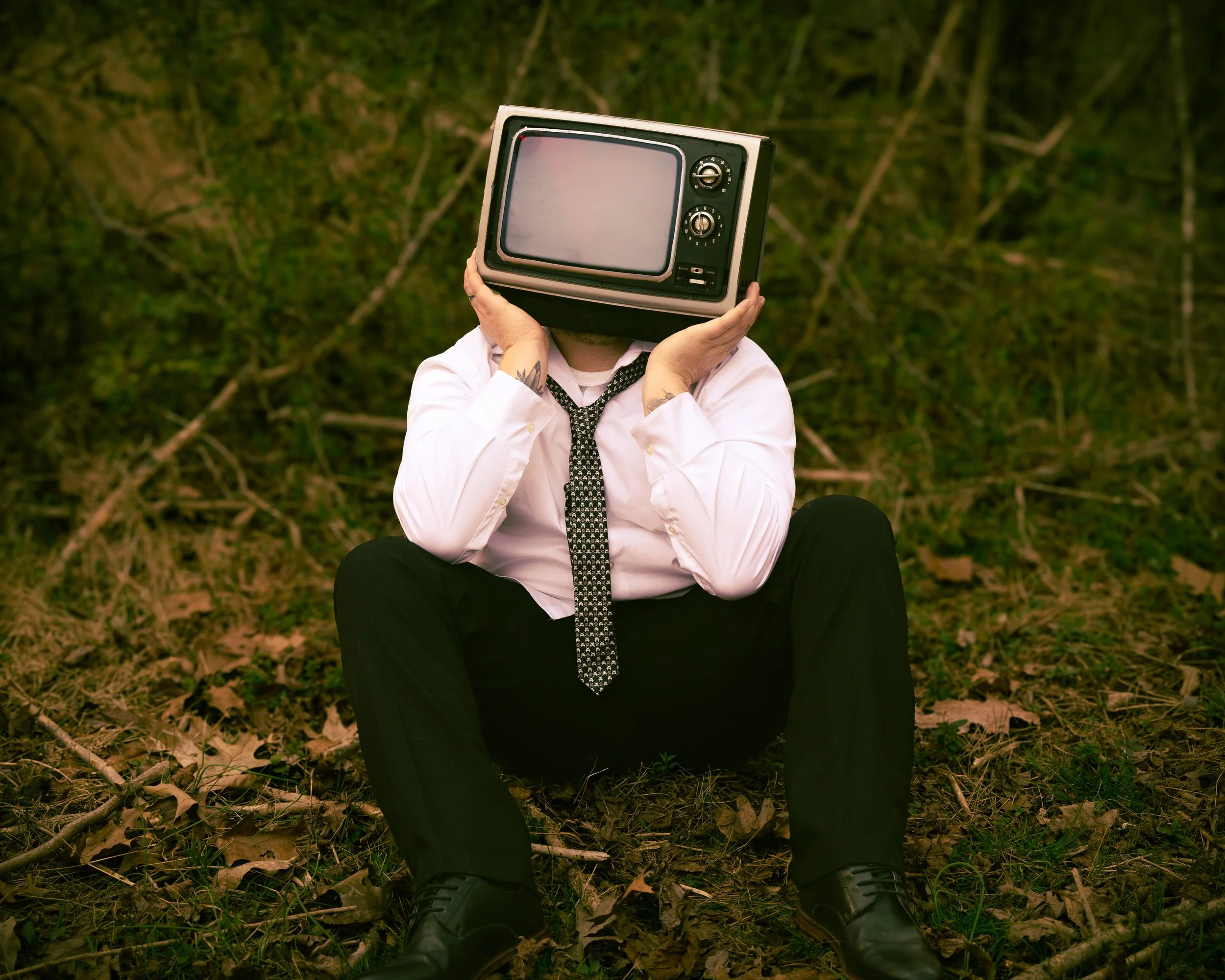 Person sitting outdoors with a vintage television set as their head, wearing a white shirt and black tie, surrounded by trees and fallen leaves.