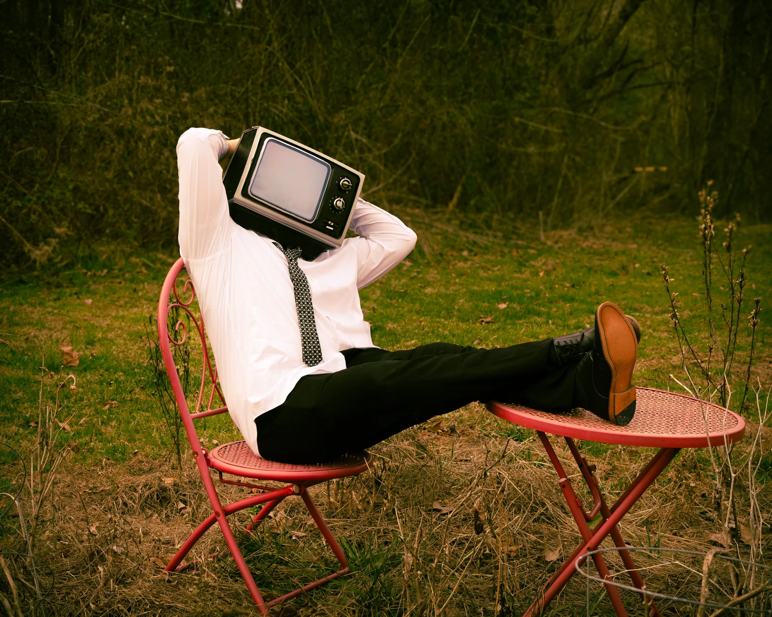 Person reclining on a pink outdoor chair in a field, wearing white shirt, black pants, and brown shoes, with a vintage television as a head, surrounded by greenery and bushes.