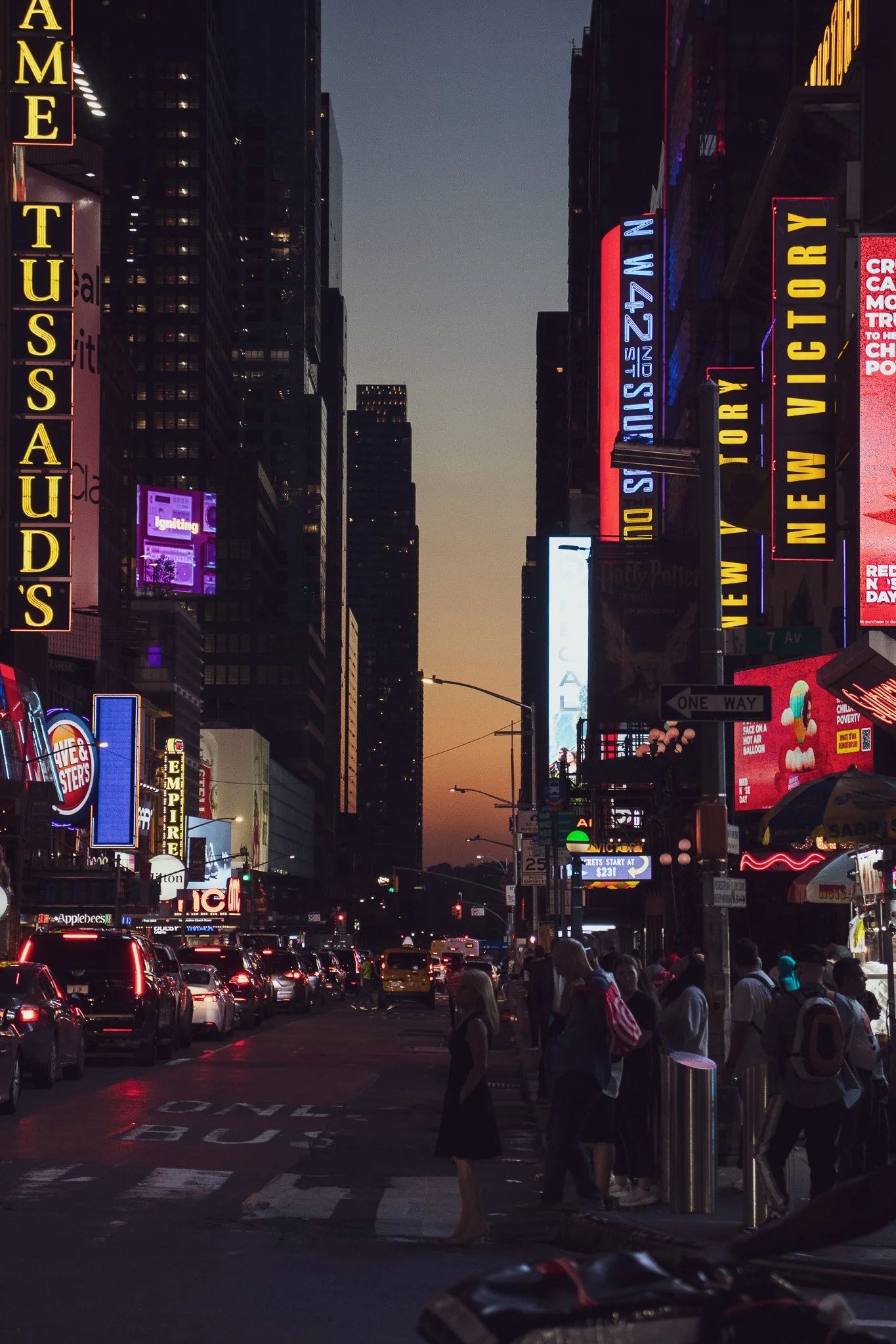 City street scene at dusk with illuminated neon signs and billboards, crowd of pedestrians crossing the street, cars stopped at traffic lights, and tall buildings in the background.