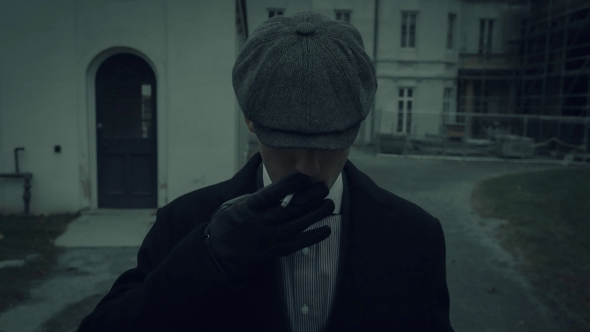 A person wearing a gray herringbone cap and black gloves, holding a cigarette near their lips, standing outdoors in front of a building.