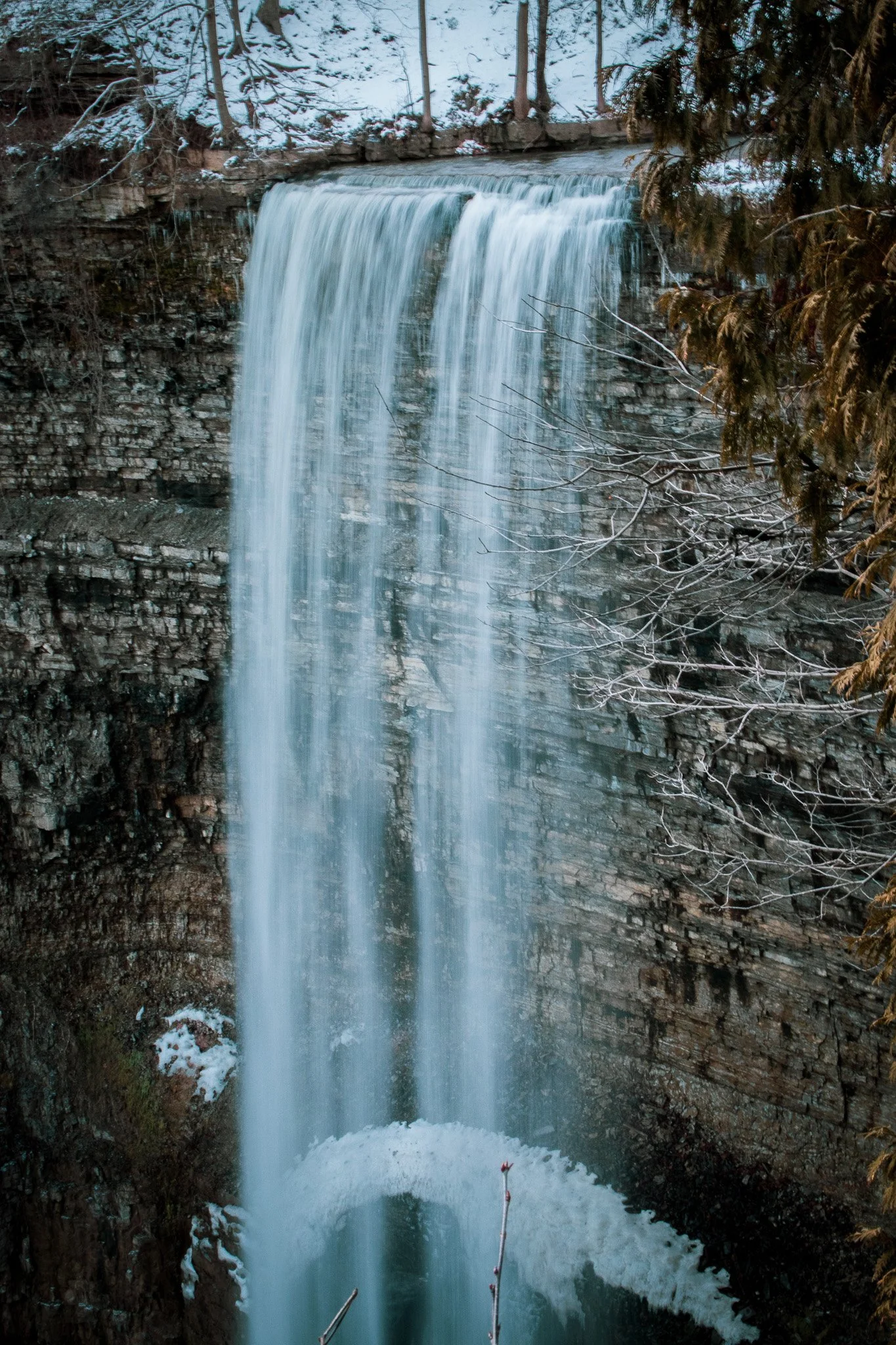 A waterfall flowing over a stone ledge in a forested area with snow and trees.