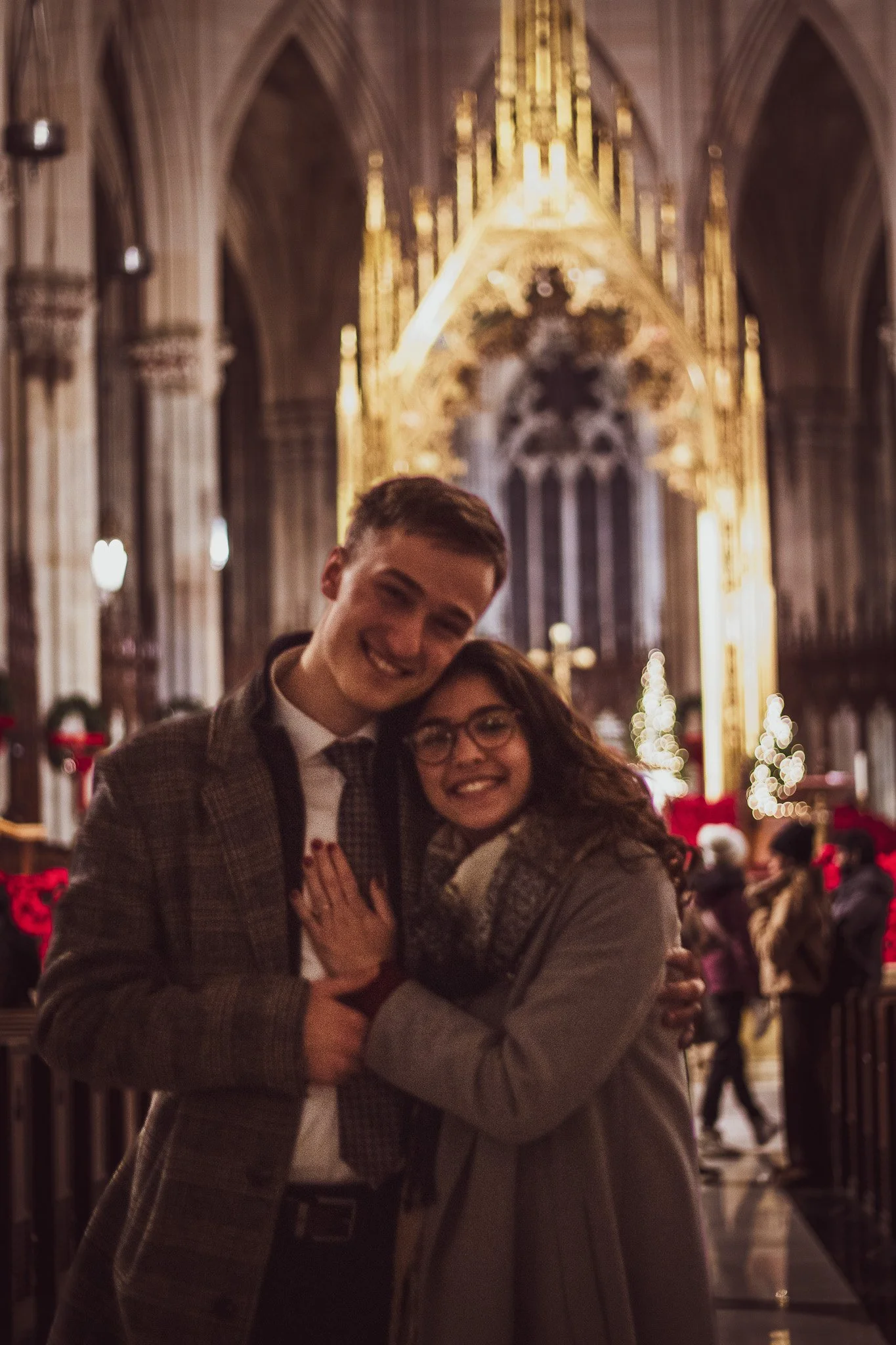 A young man and woman smiling and hugging inside a cathedral with high vaulted ceilings and Christmas lights, during the holiday season.