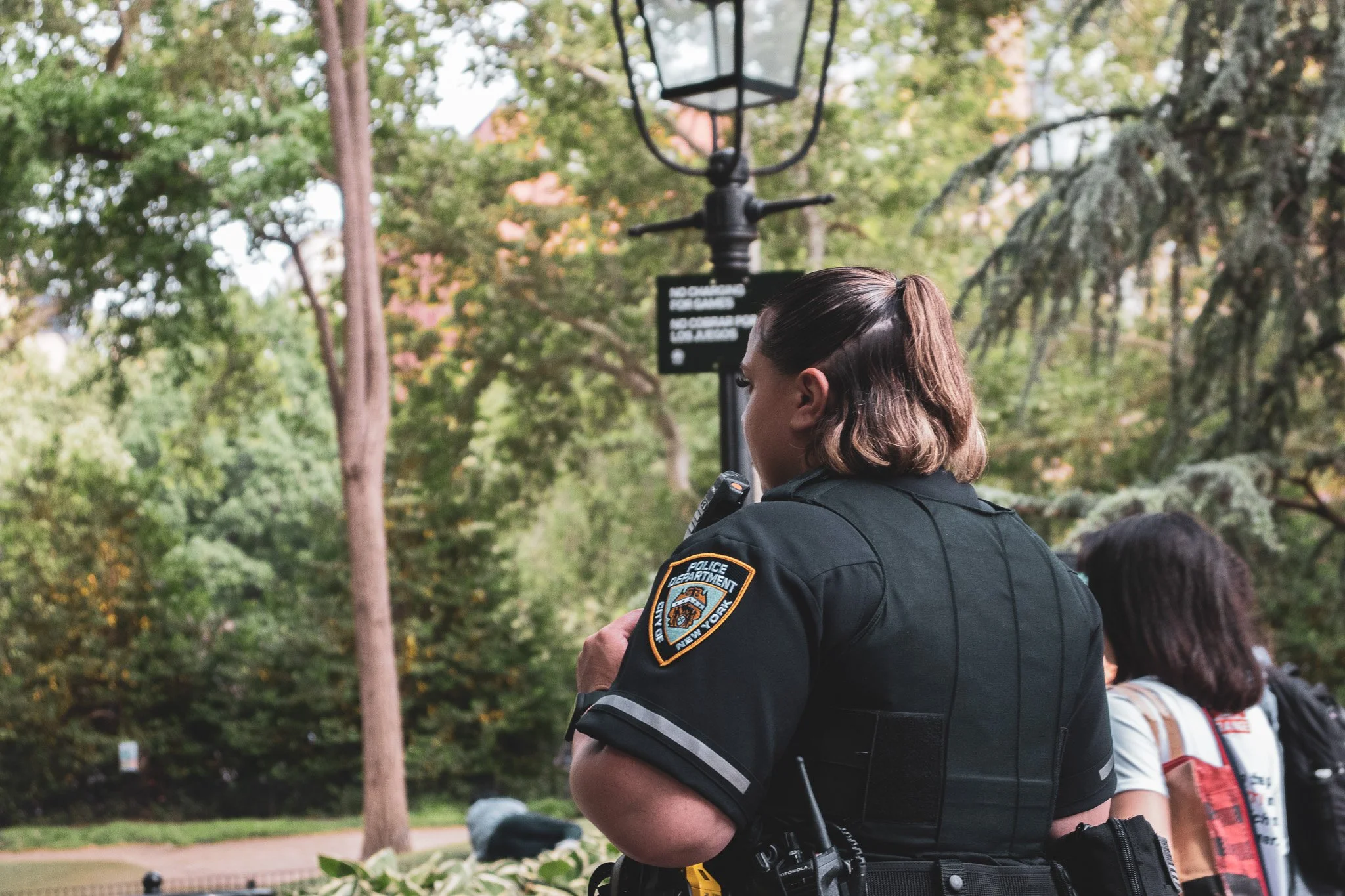A police officer standing outdoors among trees and other people, with her lower body and side profile visible. She is wearing a uniform with a patch that indicates she works for the New York City Police Department.