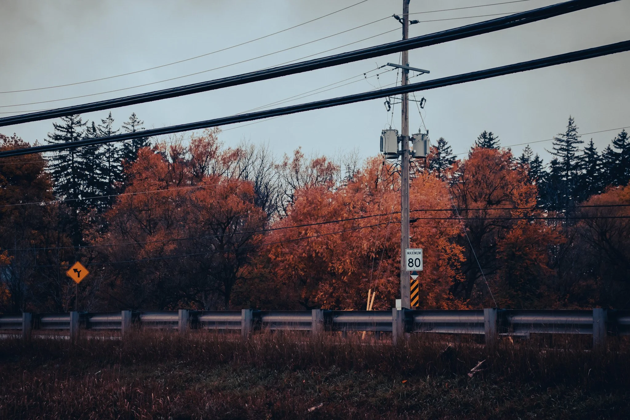 A utility pole with electrical transformers and wires in front of trees with autumn foliage, a road sign indicating a sharp turn, and a guardrail along a roadside.
