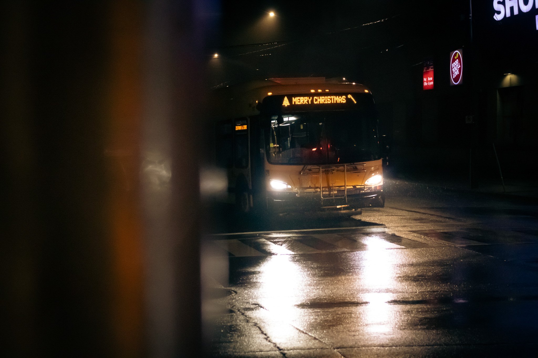 A bus with a digital sign that reads 'Merry Christmas' in the rain at night, with some illuminated signs visible on the building to the right.