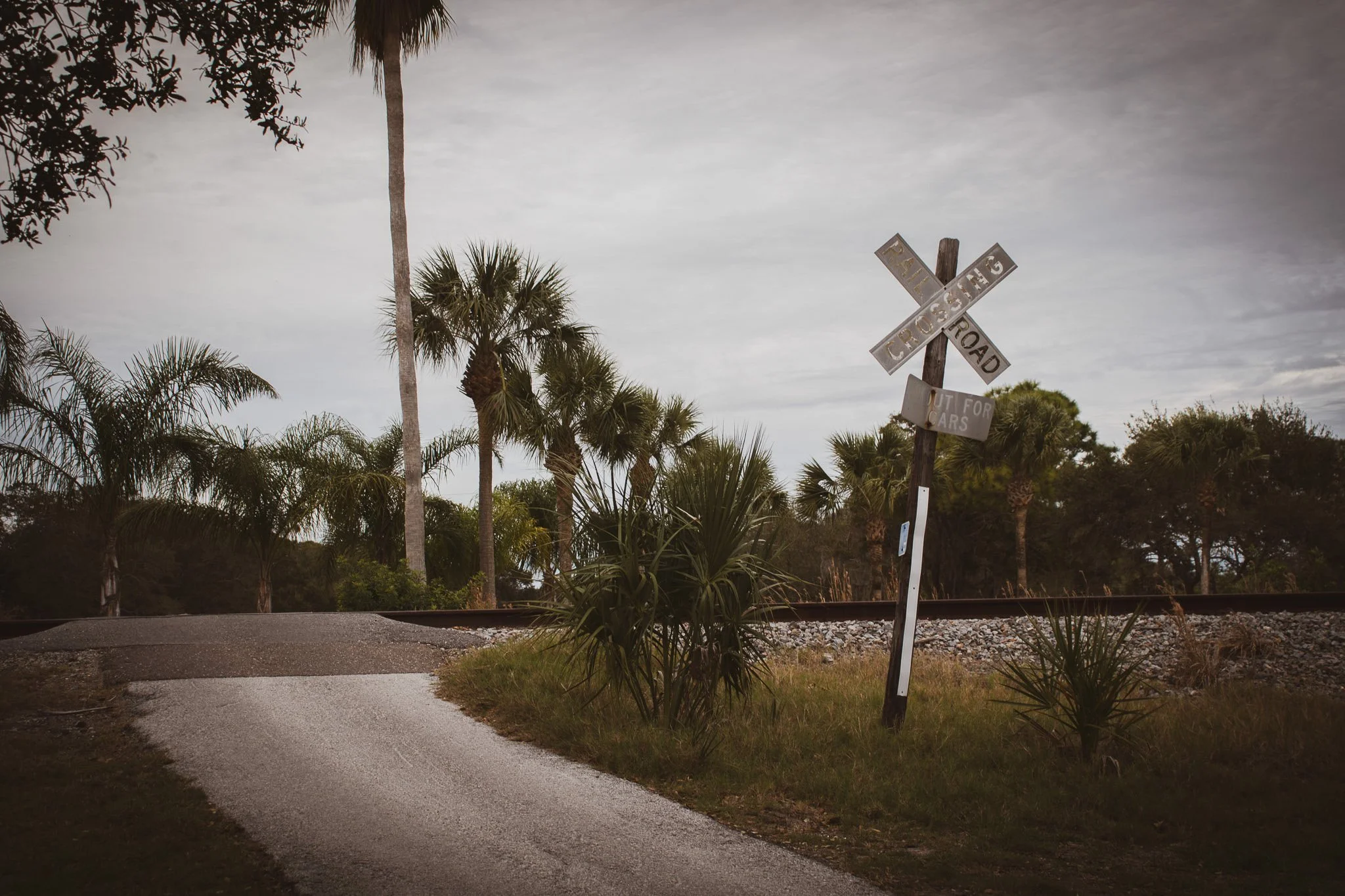 A weathered railroad crossing sign leaning to the right, situated beside a railway track with tropical trees and palm trees in the background under a cloudy sky.
