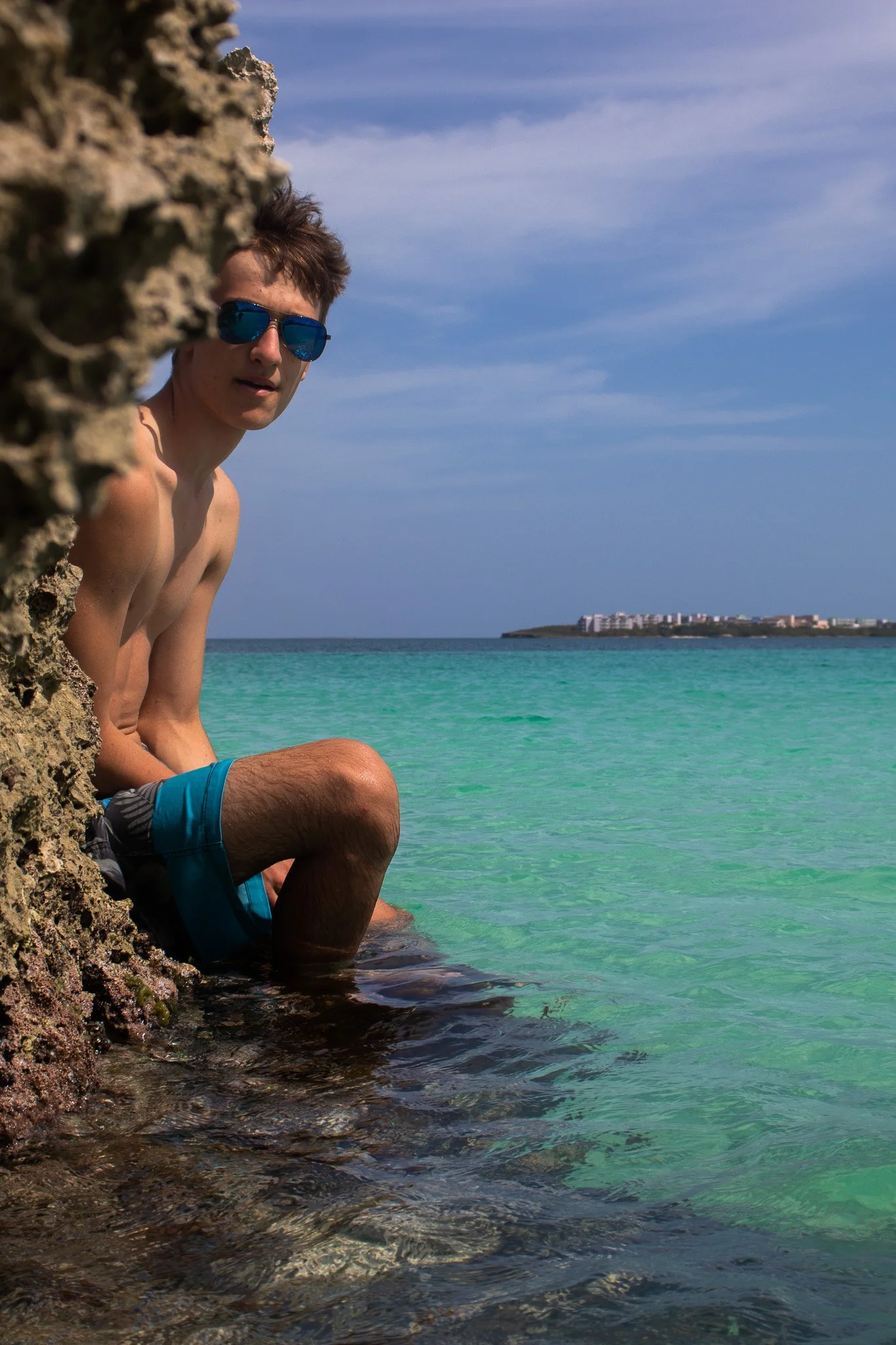A shirtless man with brown hair and blue reflective sunglasses sitting on rocks by turquoise water, with a distant landmass and buildings under a partly cloudy sky in the background.