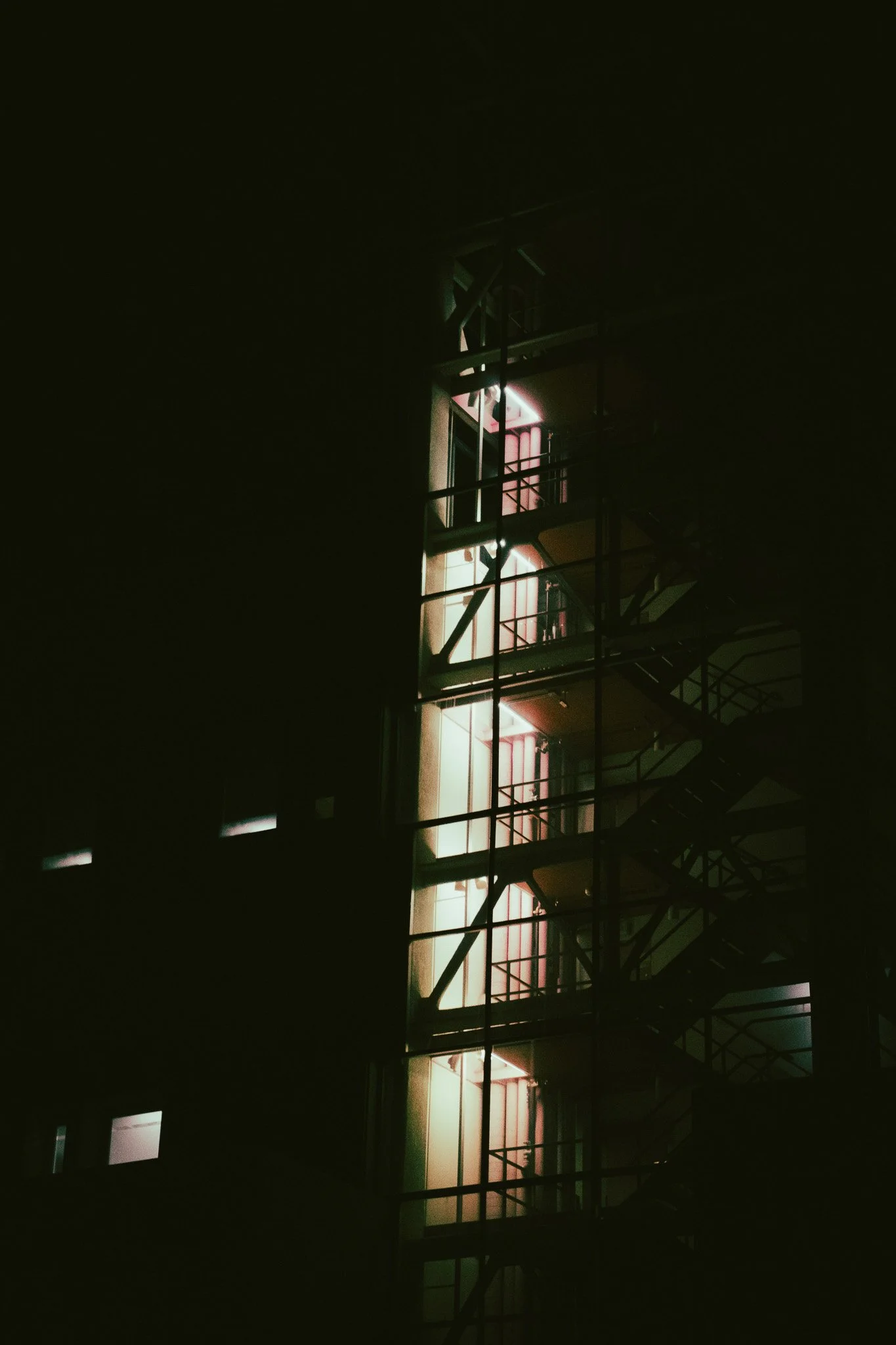 Nighttime view of a building with illuminated balconies or windows and external stairs through scaffolding or framework, with a dark sky in the background.
