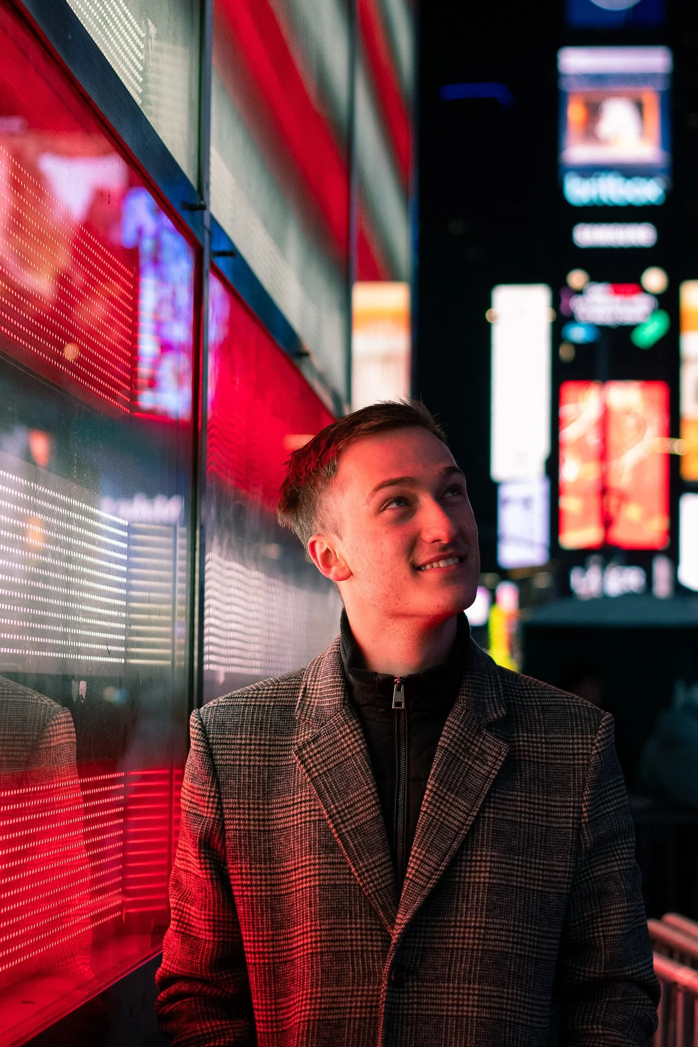 A young man wearing a plaid coat smiling in a city at night with bright neon lights and billboards in the background.