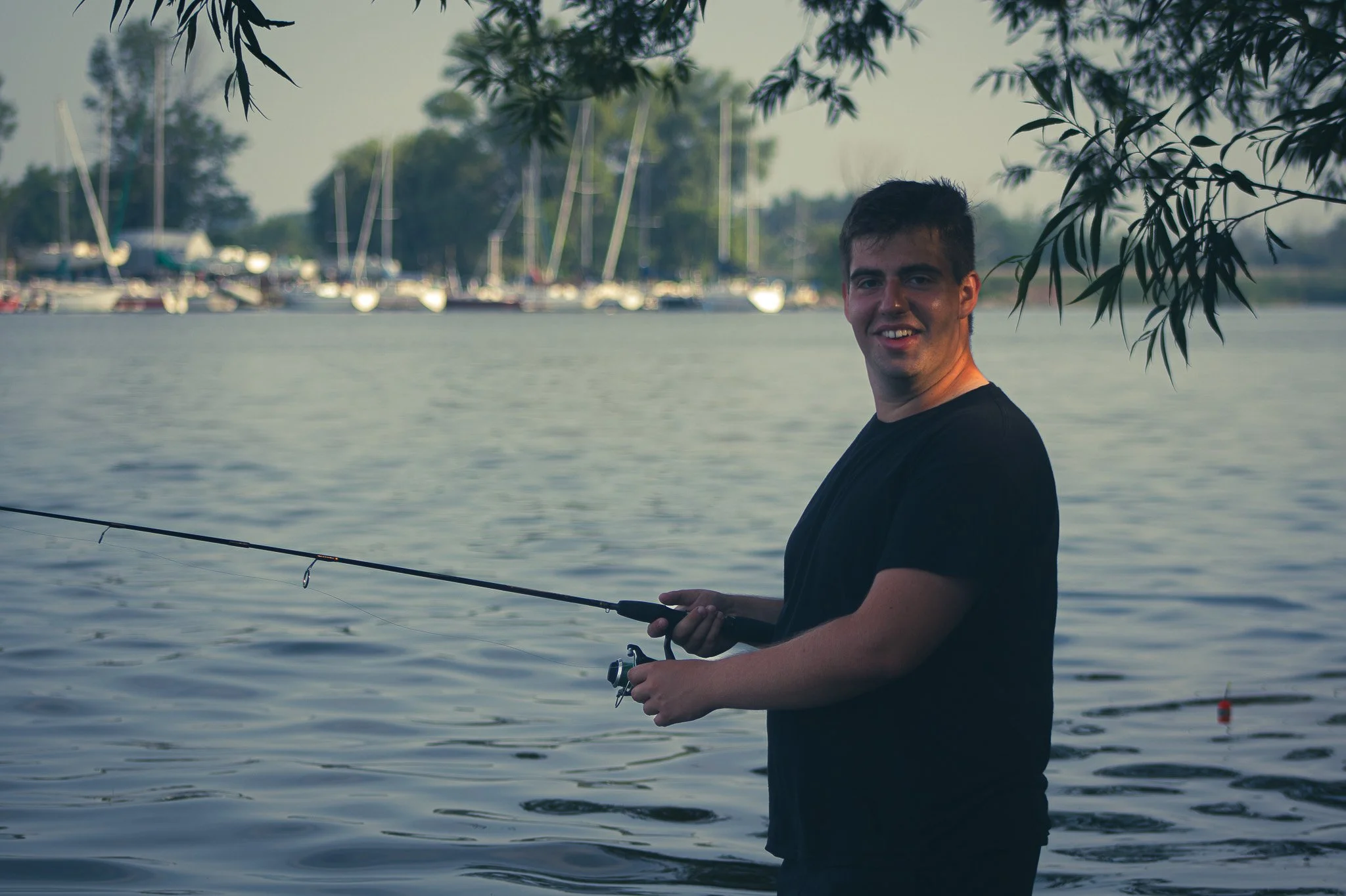 A young man fishing by a lake with sailboats anchored in the background, partially shaded by overhanging tree branches.