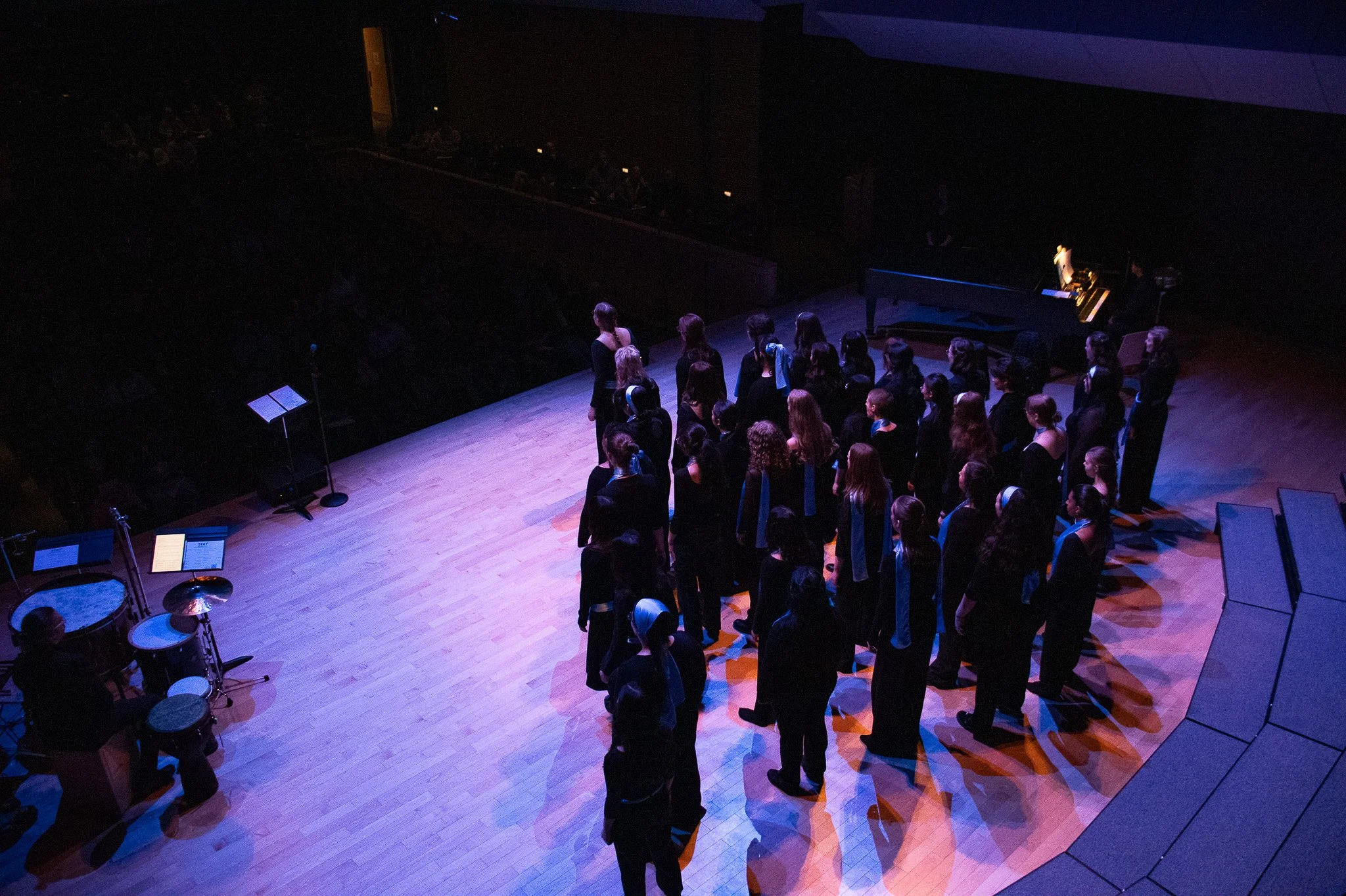 Group of people, likely choir members, standing on a stage in a dark theater with purple lighting. A grand piano is visible on the right side, and a microphone stand with sheet music is on the left. The audience is out of view.