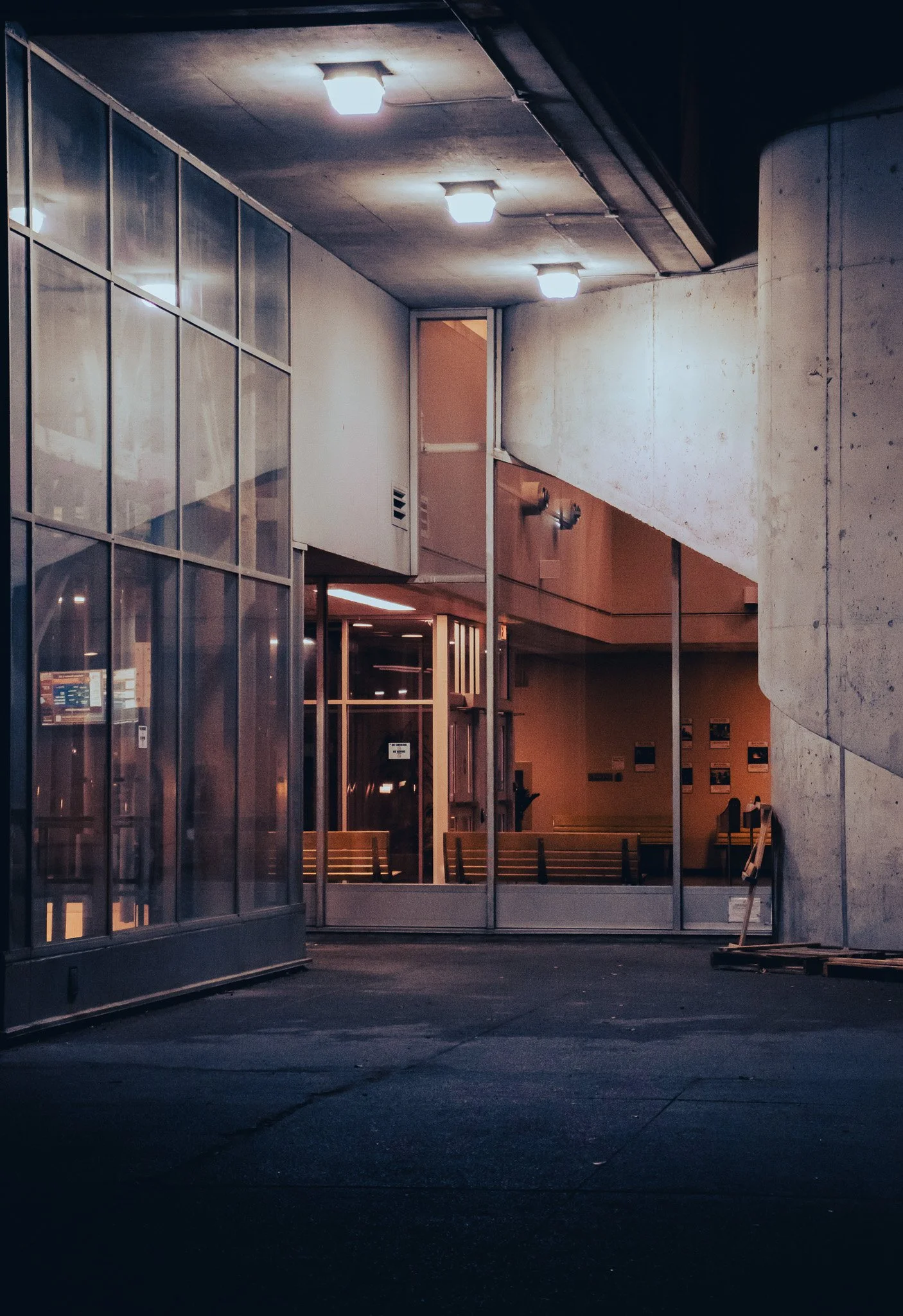 Nighttime view of an indoor area with modern lighting, glass walls, concrete columns, and benches outside.
