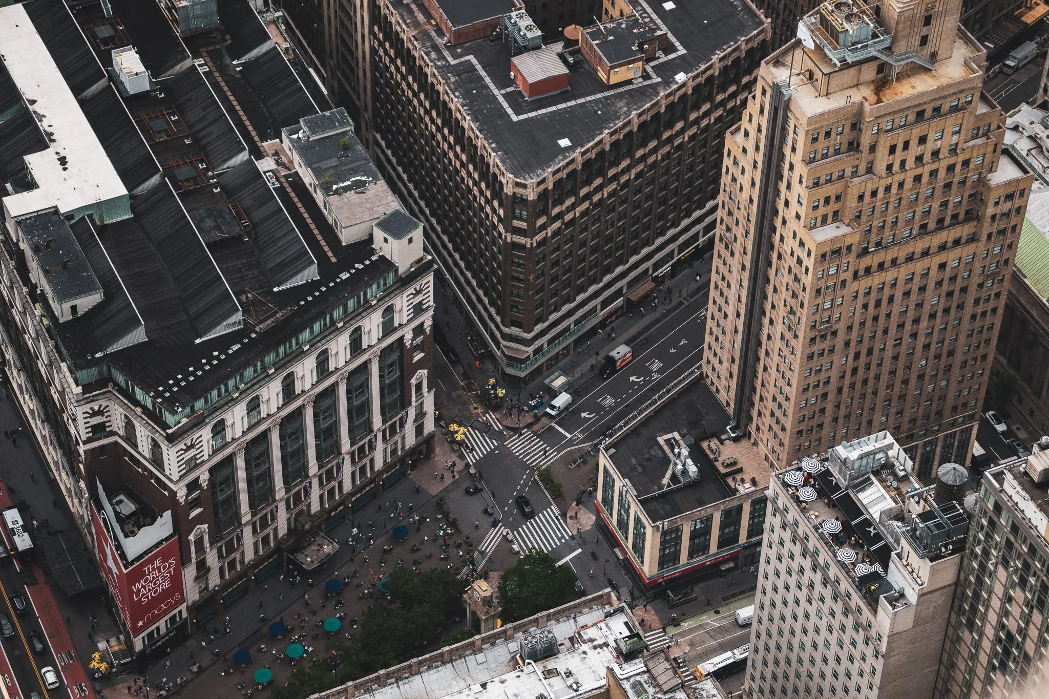 Aerial view of city buildings with streets, pedestrians, and vehicles, including Macy's store with a red sign, and sidewalks with umbrellas at street level.