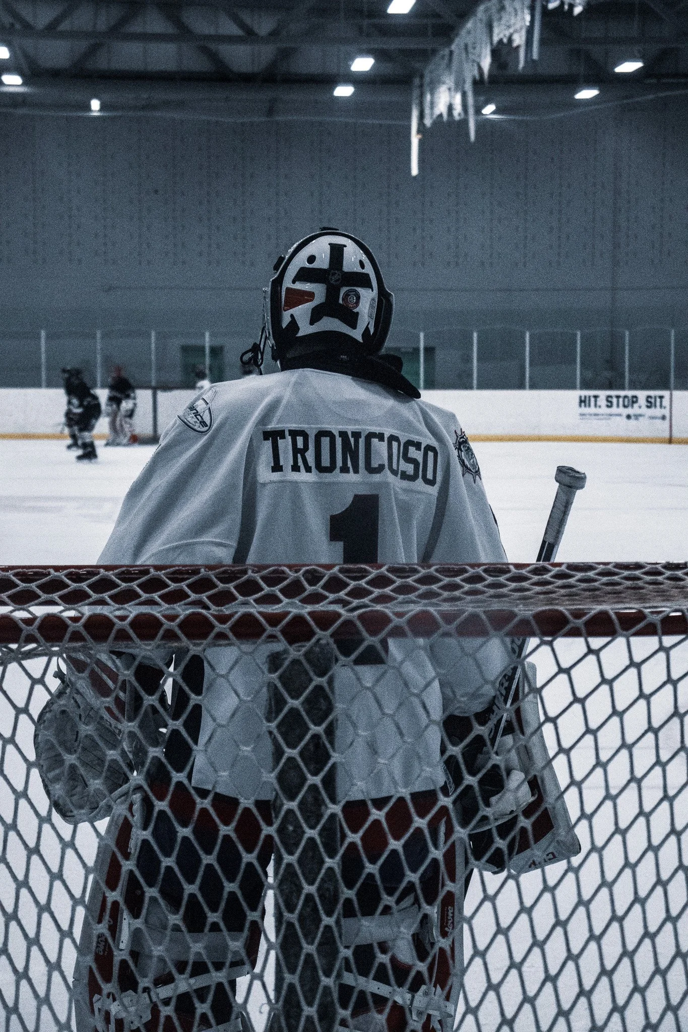 Ice hockey goalie in full gear stands in front of the net, facing the ice rink with other players in the background.