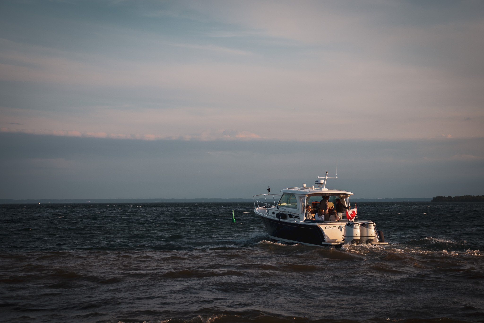 A white boat with a mechanical design on the water with people onboard, with the name 'Salty' visible on the side, surrounded by a large body of water under a mostly cloudy sky.