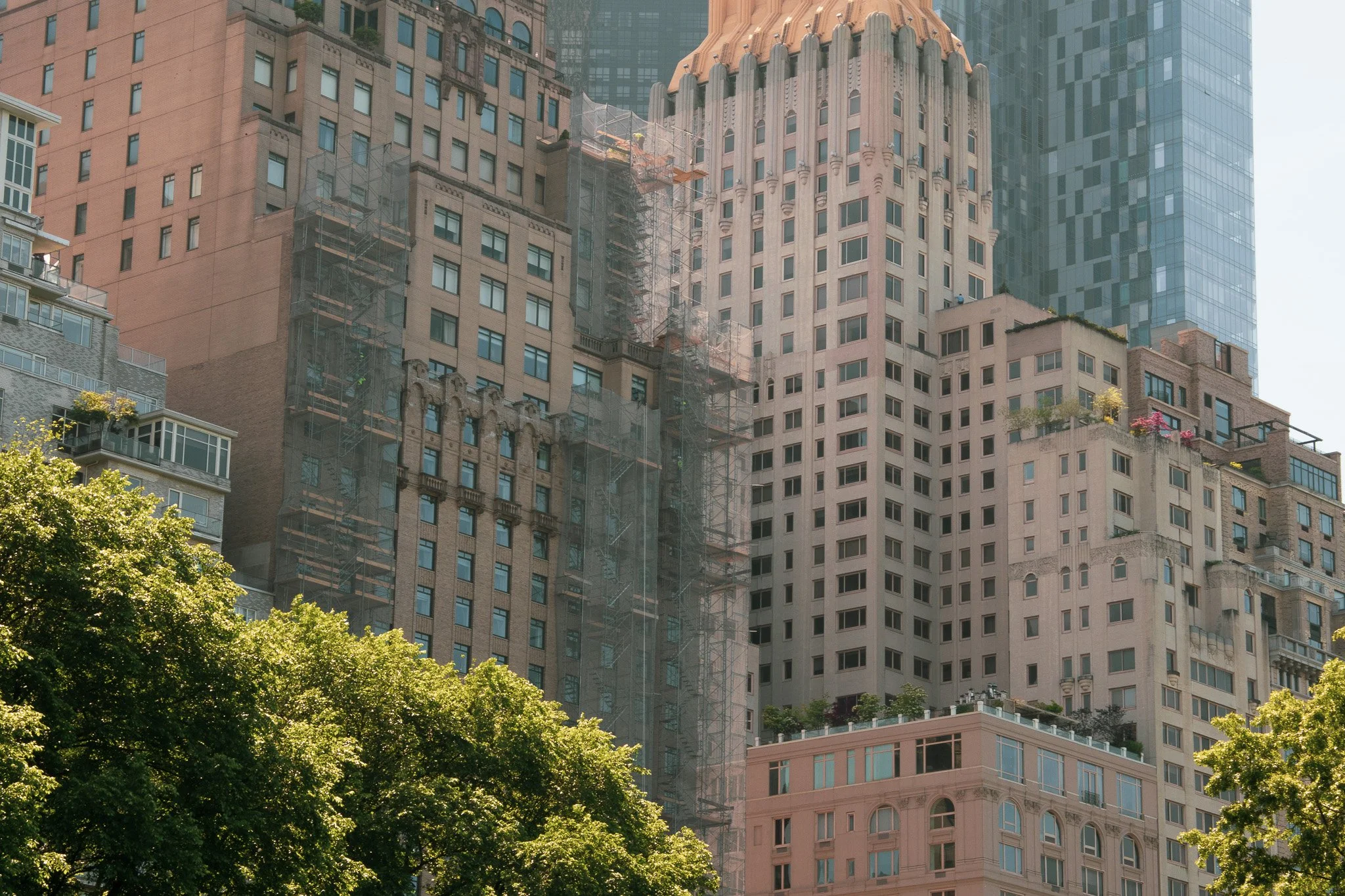 Tall city buildings, some under construction with scaffolding, and lush green trees at the bottom in an urban setting.