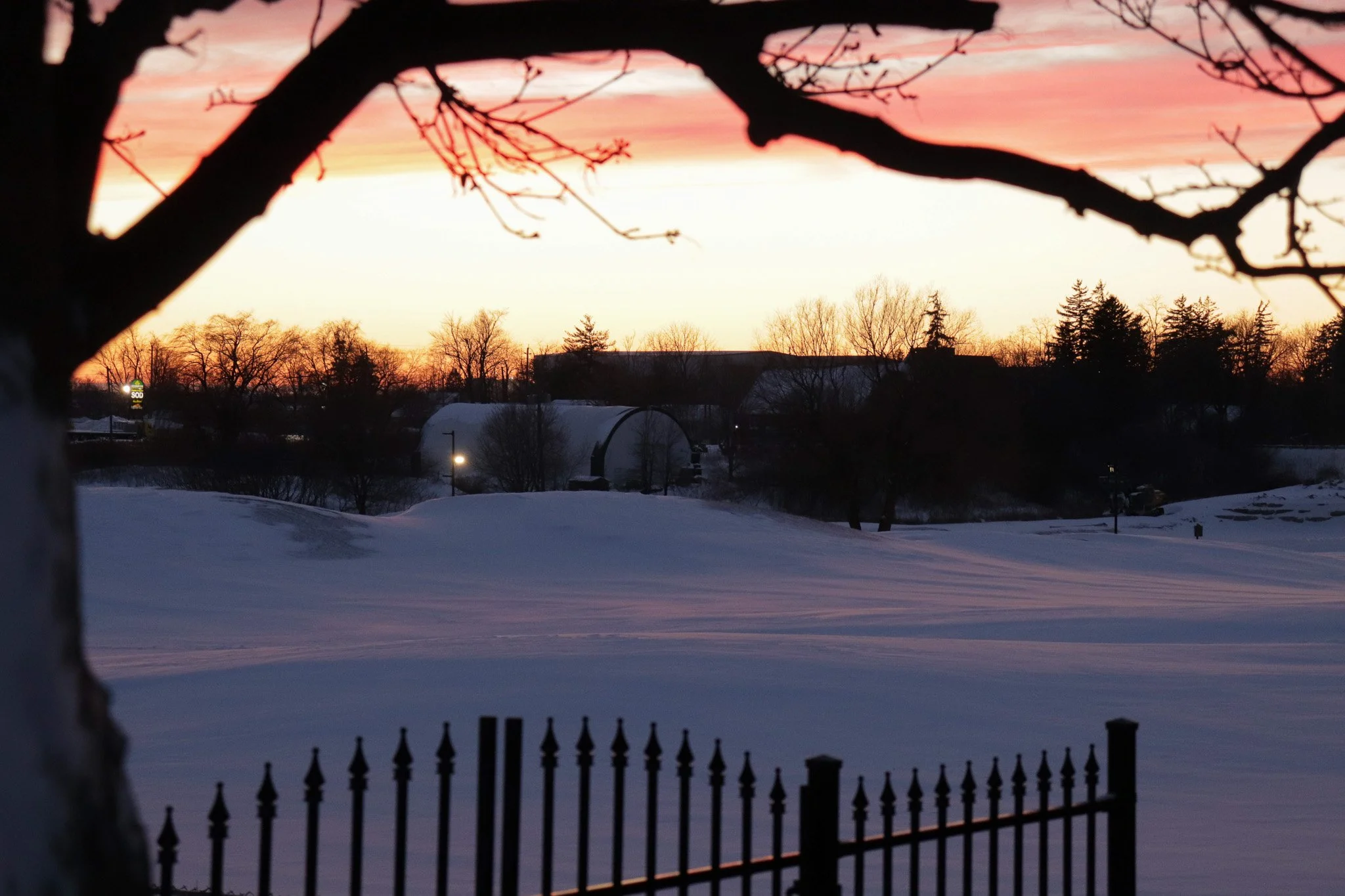 Sunset view with colorful orange and pink sky, scattered trees, snowy landscape, and a black metal fence in the foreground.