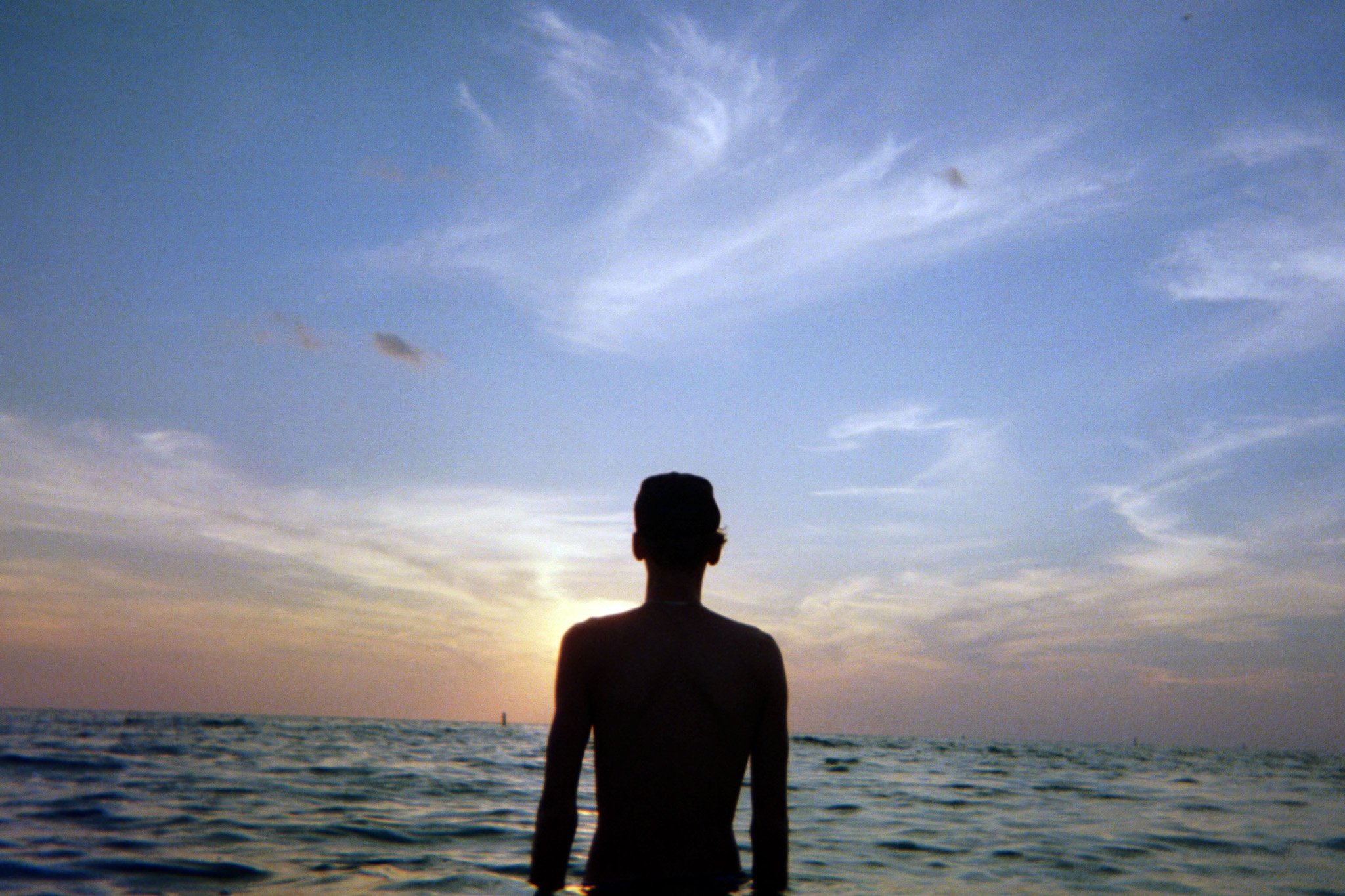 A person standing on the beach facing the ocean during sunset, with a sky filled with wispy clouds.