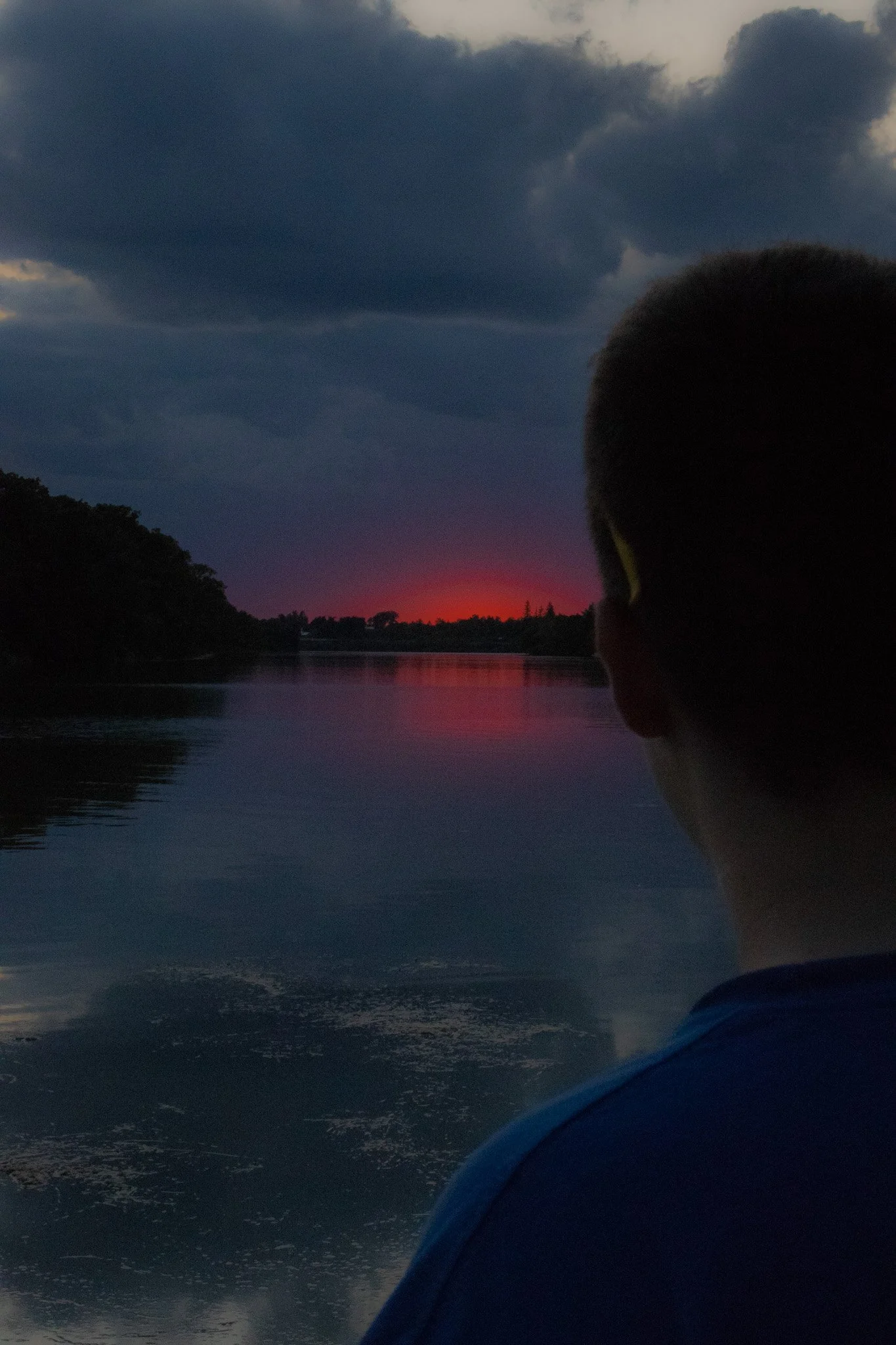 A person looking out over a calm body of water during sunset with dark clouds in the sky, and trees on the horizon.
