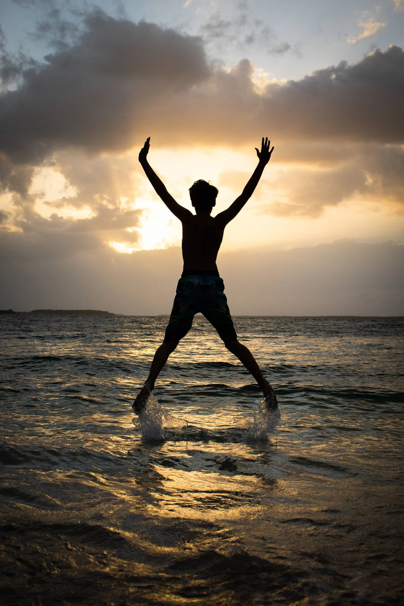 A silhouette of a person jumping in the water at sunset with arms raised, with cloudy sky and horizon in the background.