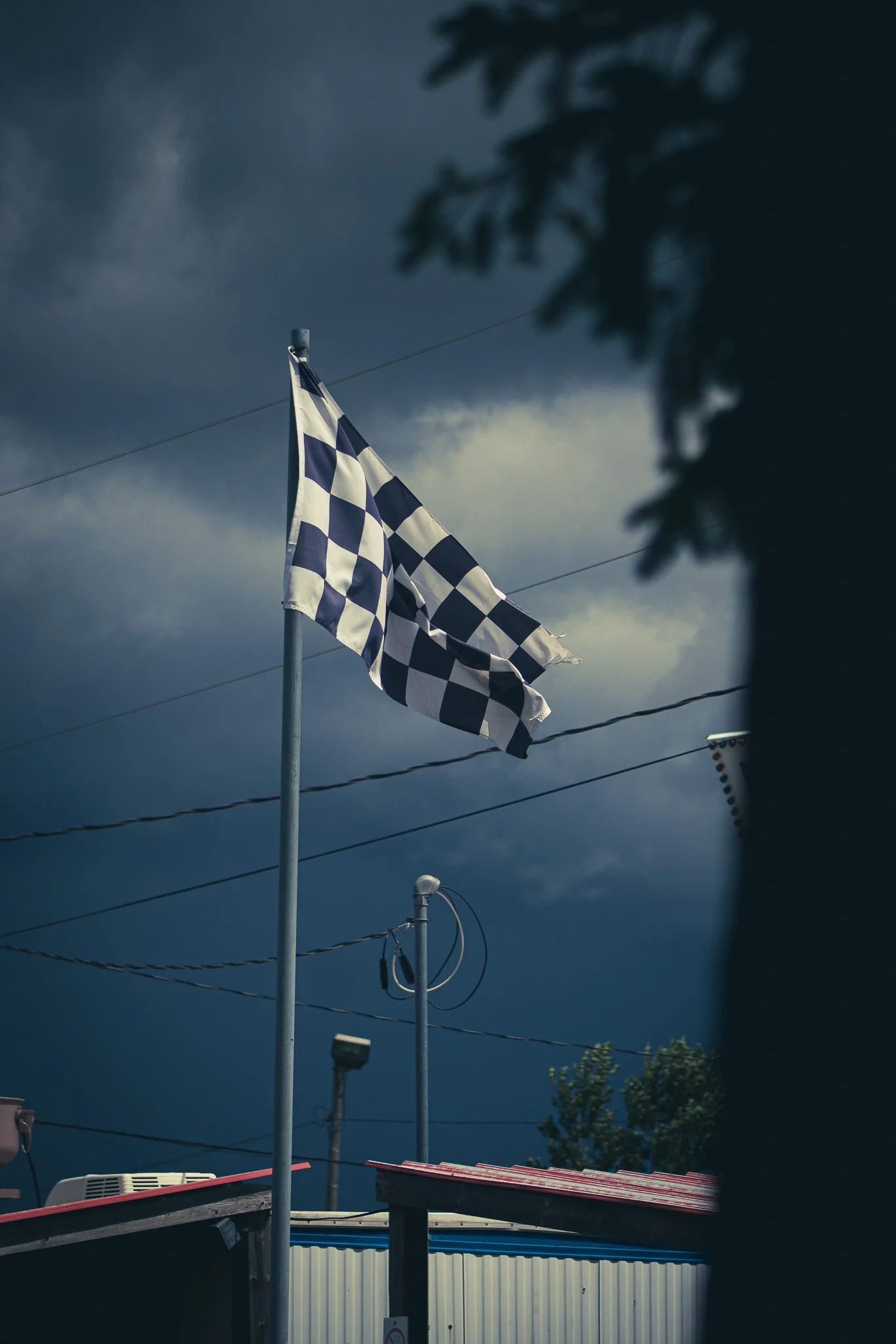 Dark cloudy sky with a checkered flag on a pole, surrounded by power lines and trees in the background.