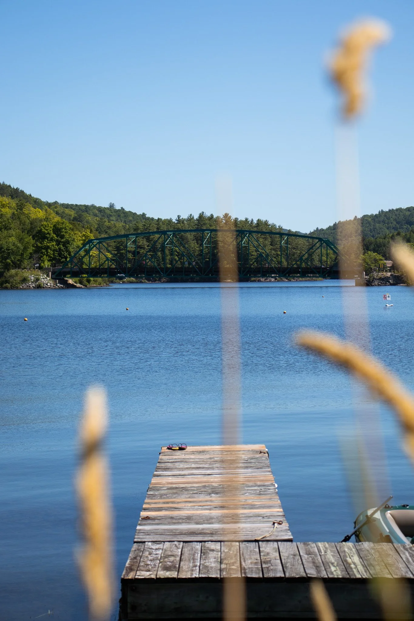 A wooden dock extends into a lake with a small boat docked to the right side. In the background, a green metal bridge crosses the lake, surrounded by green trees and hills. The sky is clear and blue.