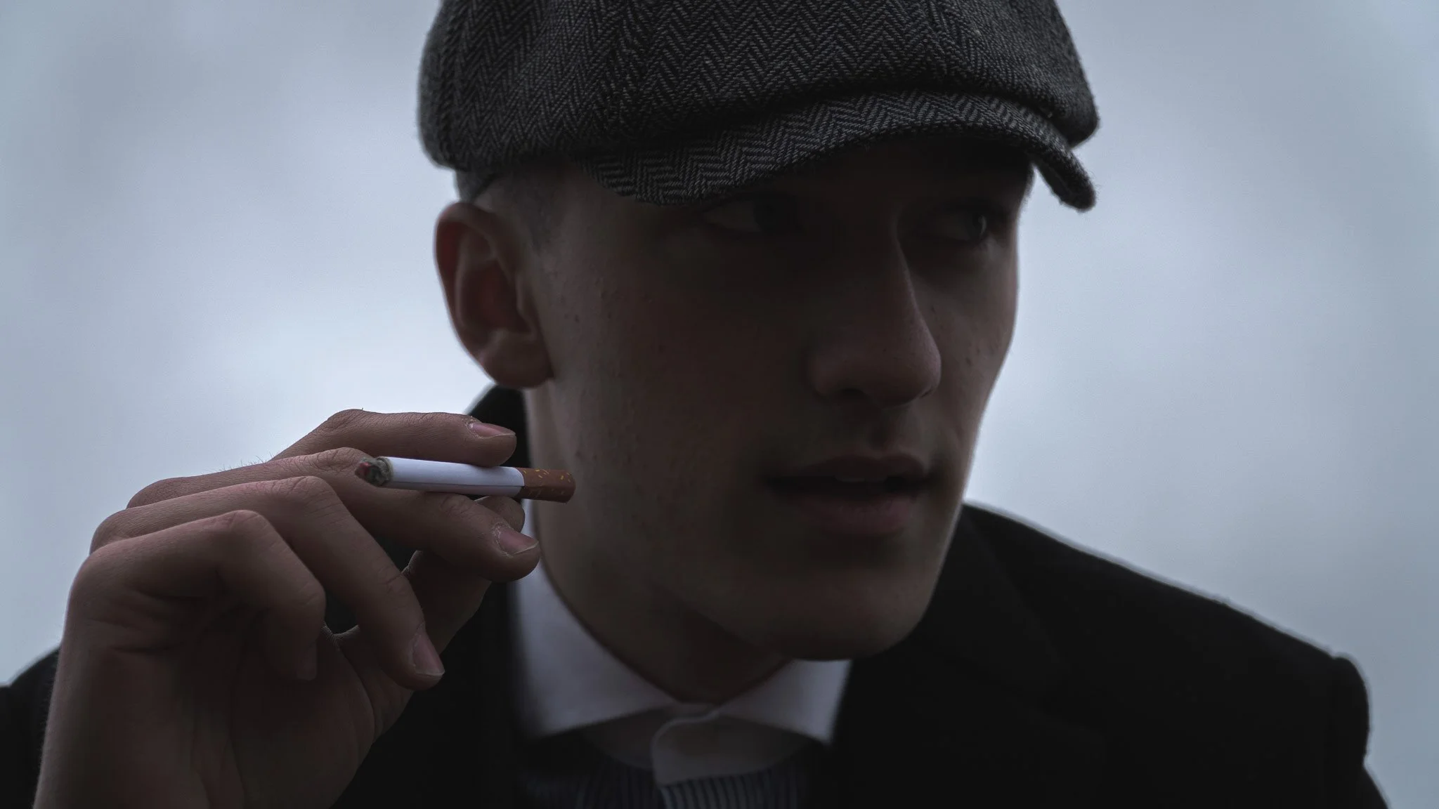 Close-up of a young man wearing a gray cap and dark jacket, holding a cigarette near his mouth, with a serious expression in a dim, overcast setting.