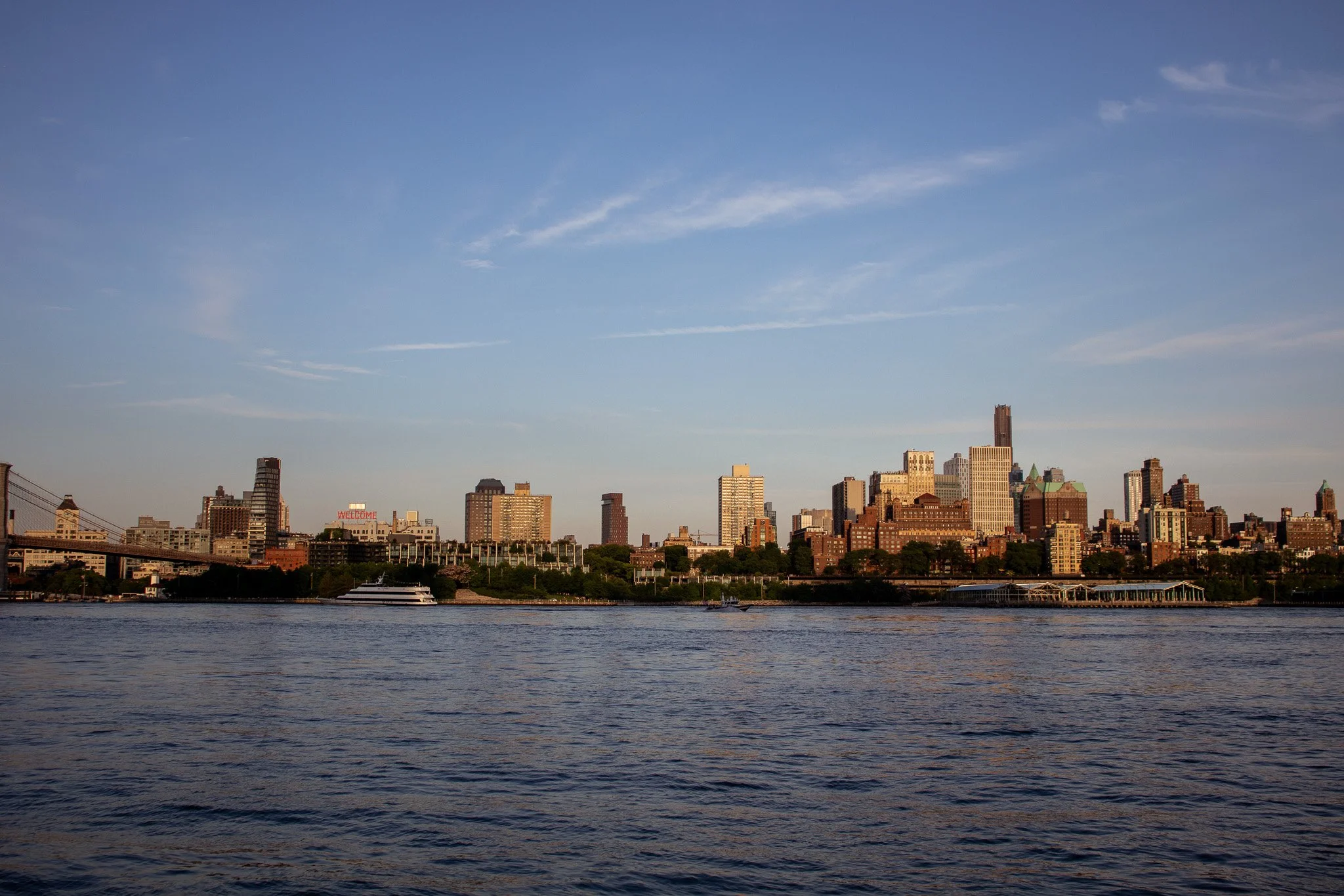 View of a city skyline across a body of water, with various tall buildings and a bridge on the left, under a blue sky with a few clouds.
