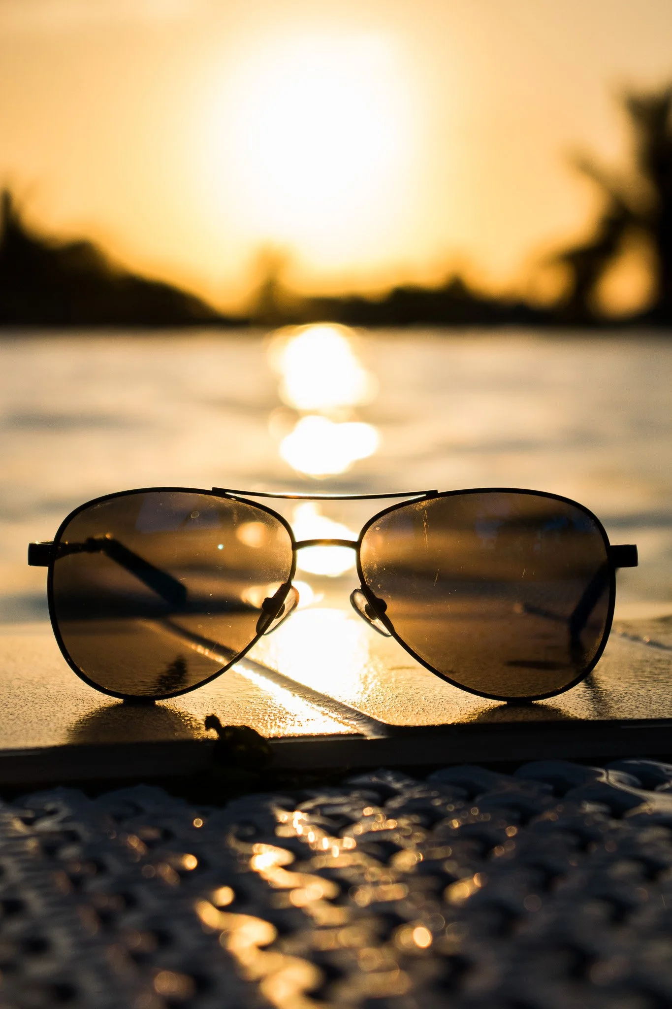 Sunglasses placed on a textured surface with a sunset over water and trees in the background.