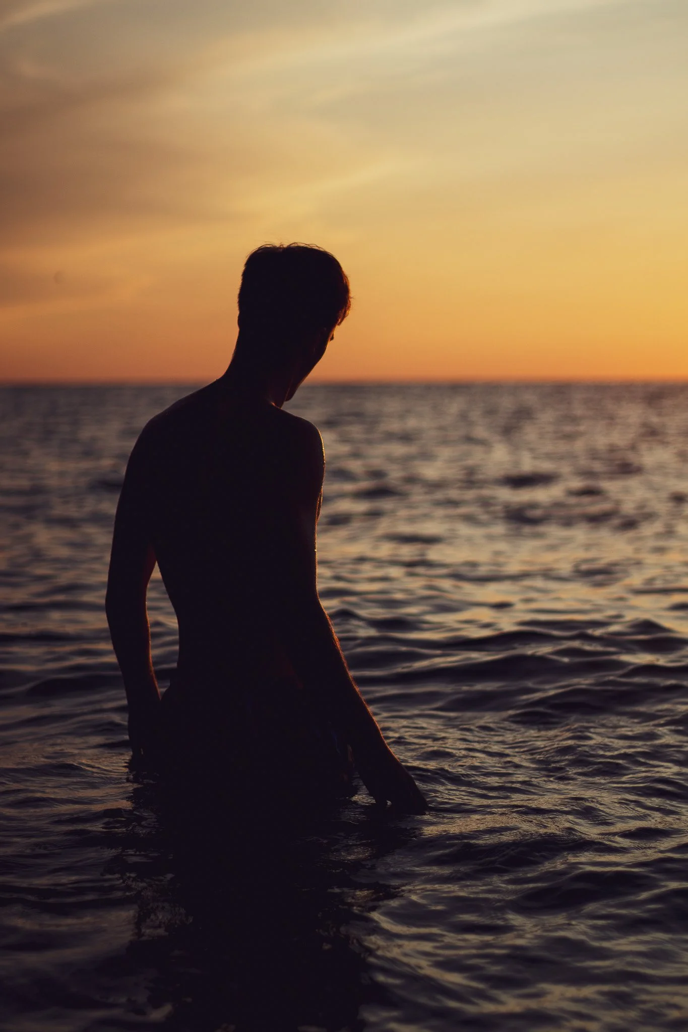 Silhouette of a person sitting by the water during sunset, with the sky painted in shades of orange and yellow.