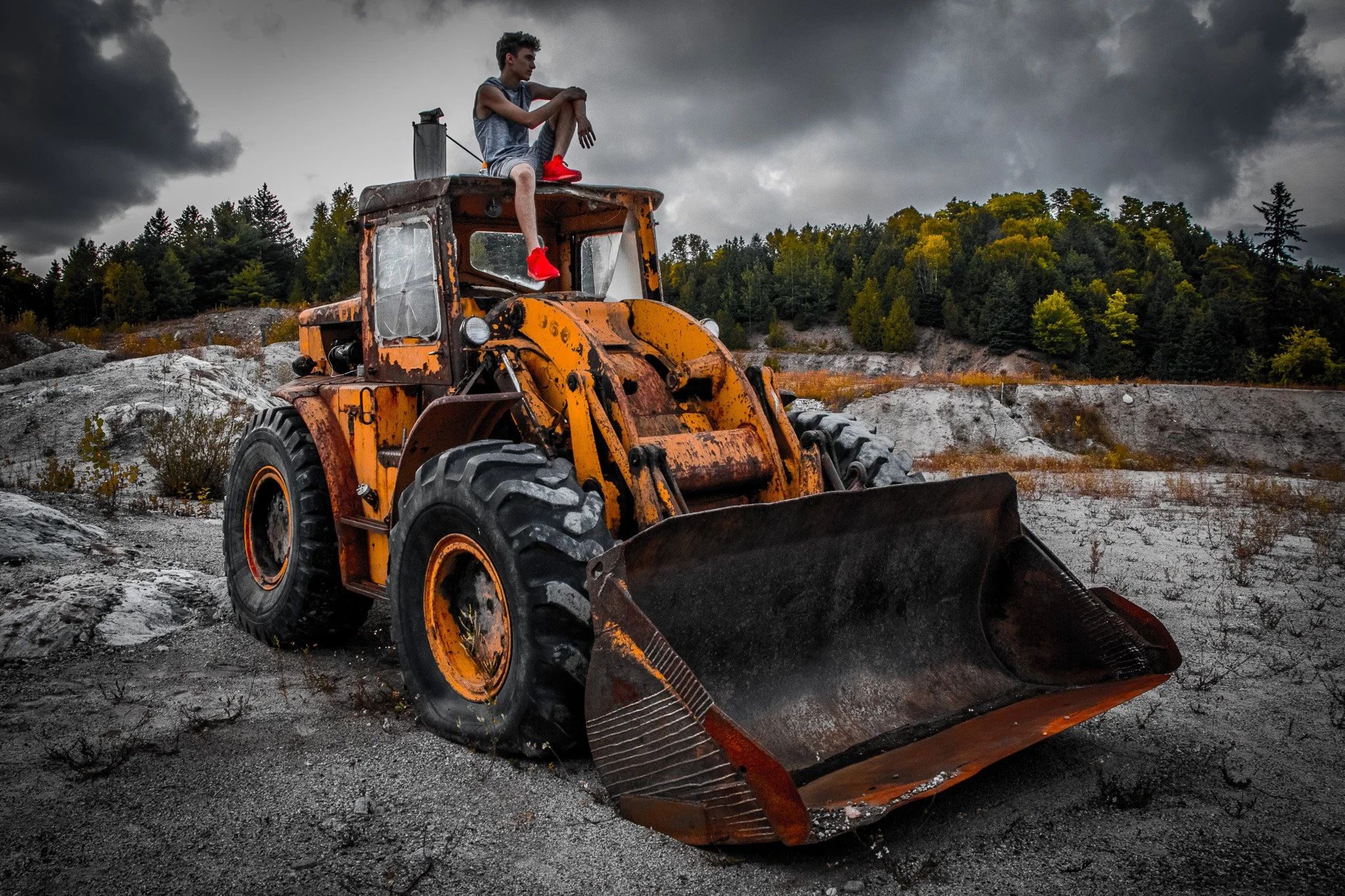 A young man sitting on top of a rusty excavator in a quarry with dark stormy clouds overhead and a forested hillside in the background.
