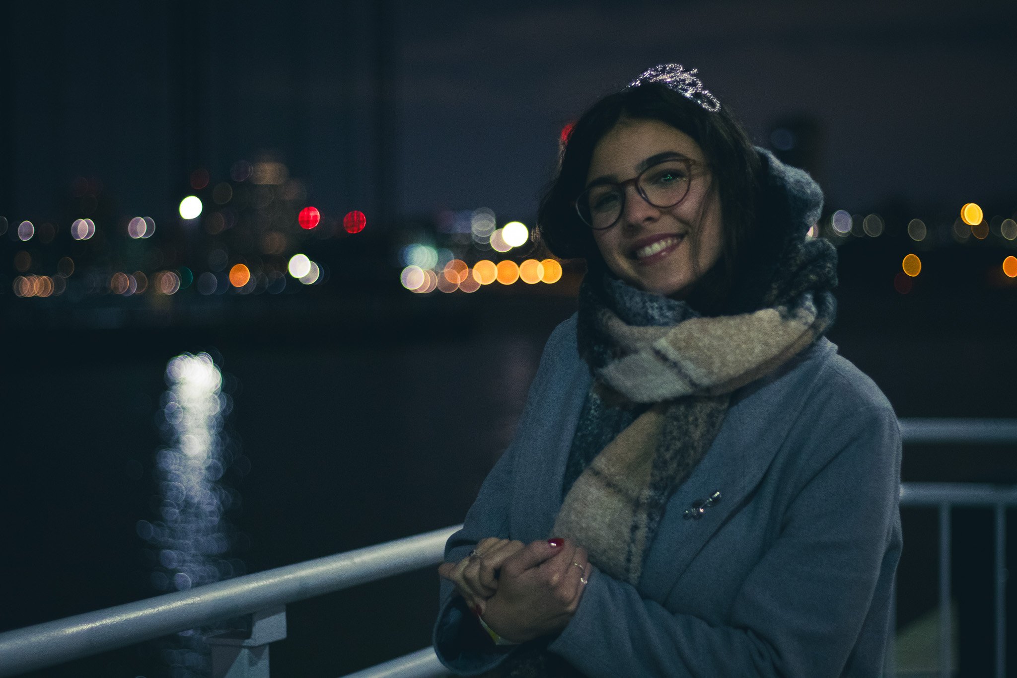 A woman with glasses, a scarf, and a gray coat standing outdoors at night near a body of water with city lights in the background.