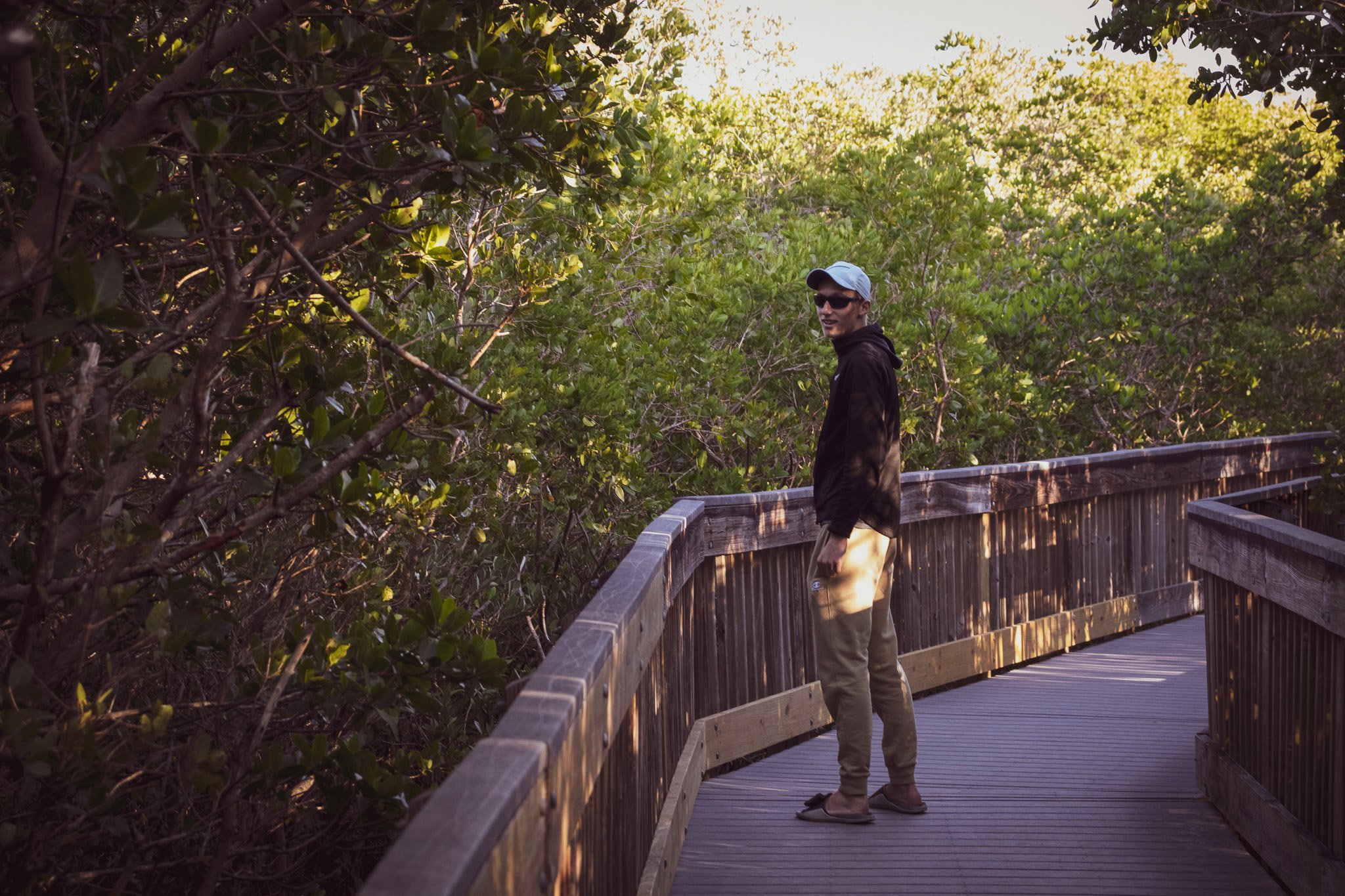 A man with a light-colored cap, dark sunglasses, a black hoodie, and beige pants standing on a wooden boardwalk surrounded by green shrubbery at sunset.