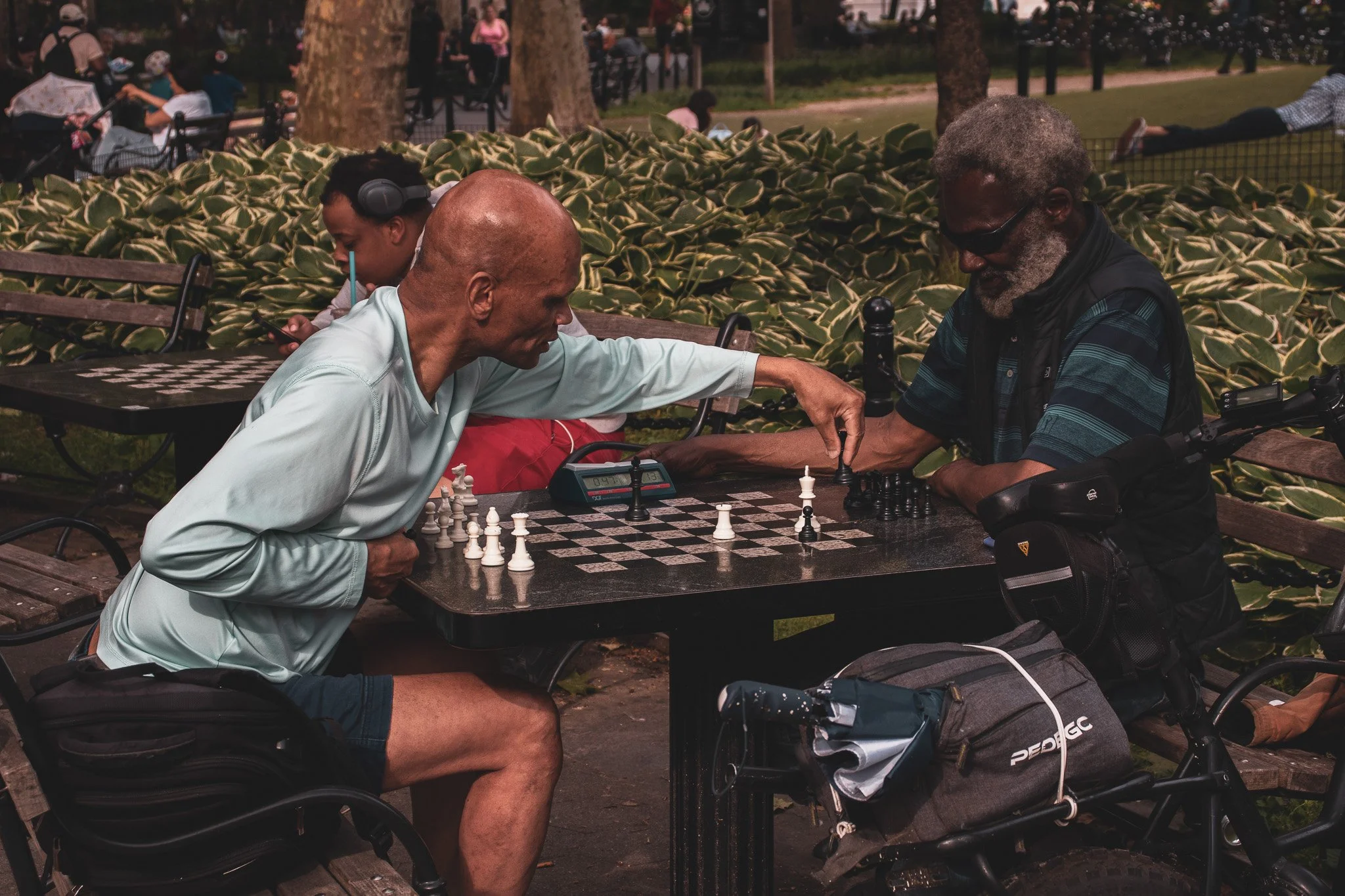 Three people are engaged in a game of chess outdoors at a park. Two men are at the table, one 's playing chess and the other is looking at a smartphone. The third person, a young woman, is sitting nearby with headphones on. They are surrounded by gre