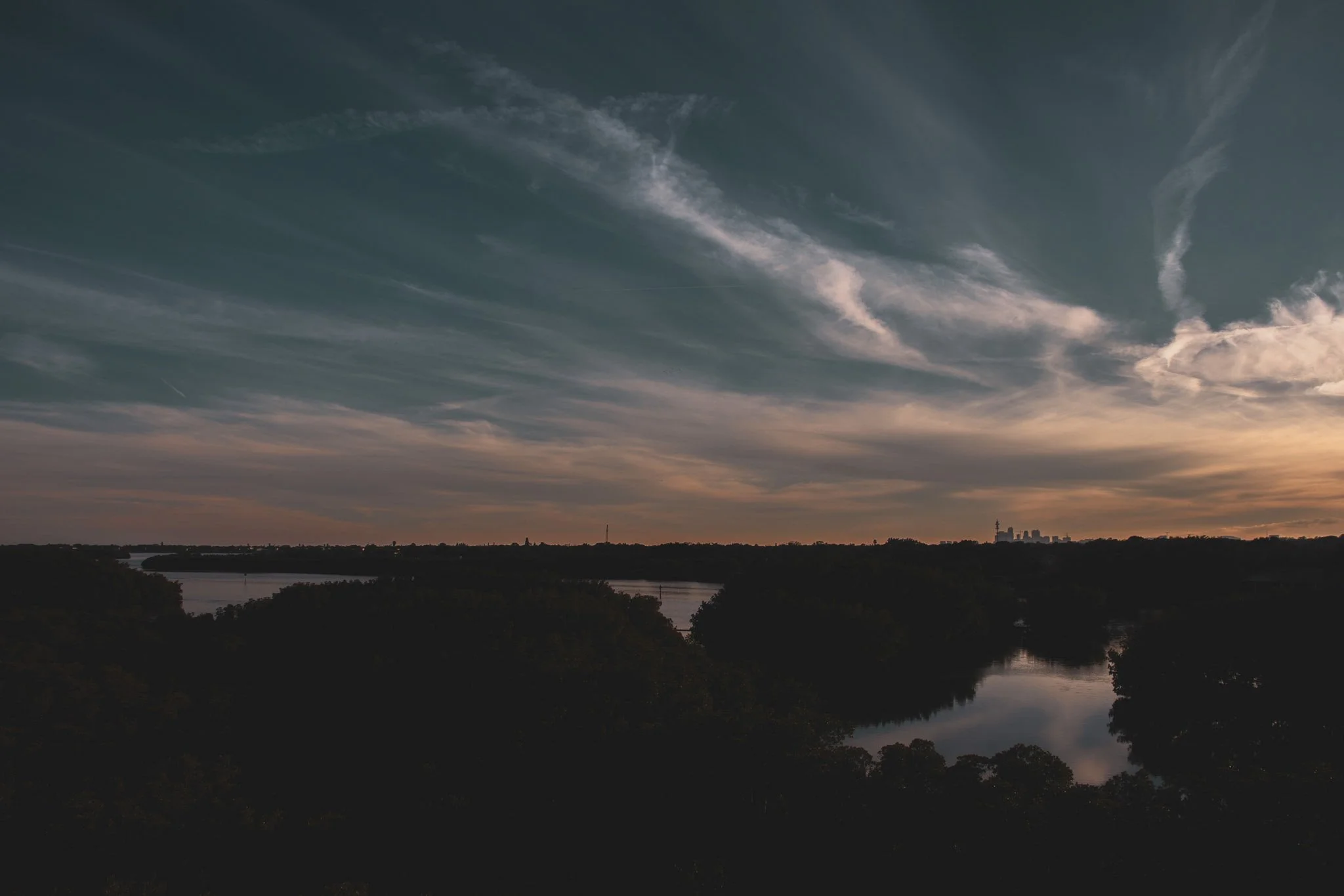 A photo of a scenic river view at sunset with a dark silhouette of trees and buildings in the distance, and a sky with streaks of clouds.