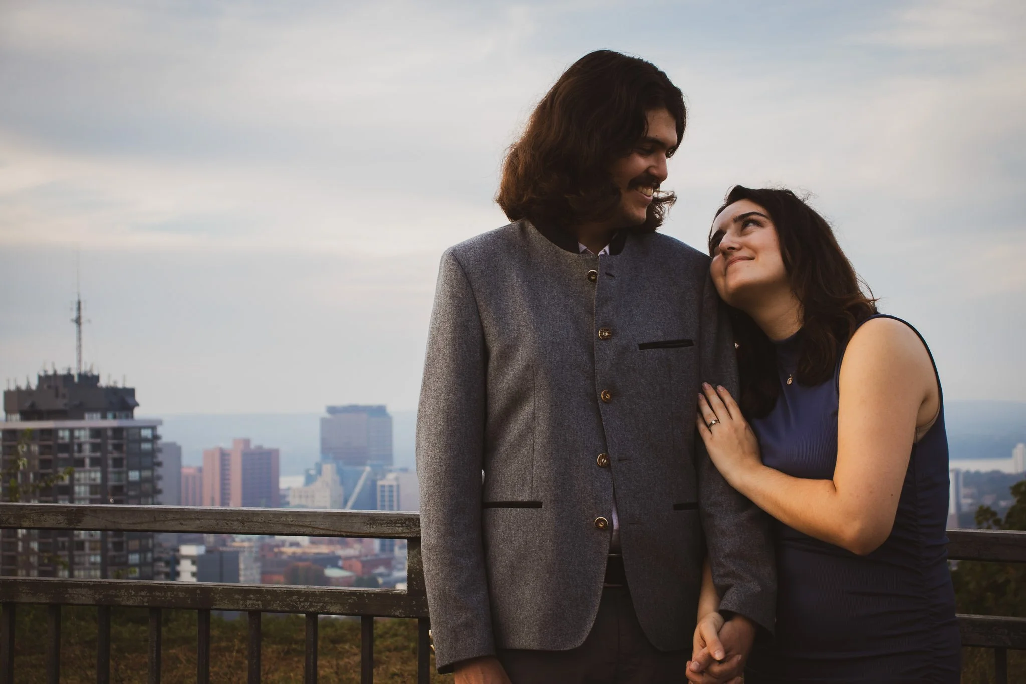 A man and woman share a tender moment on a city rooftop with a skyline view, smiling at each other, the woman leaning her head on the man's chest.