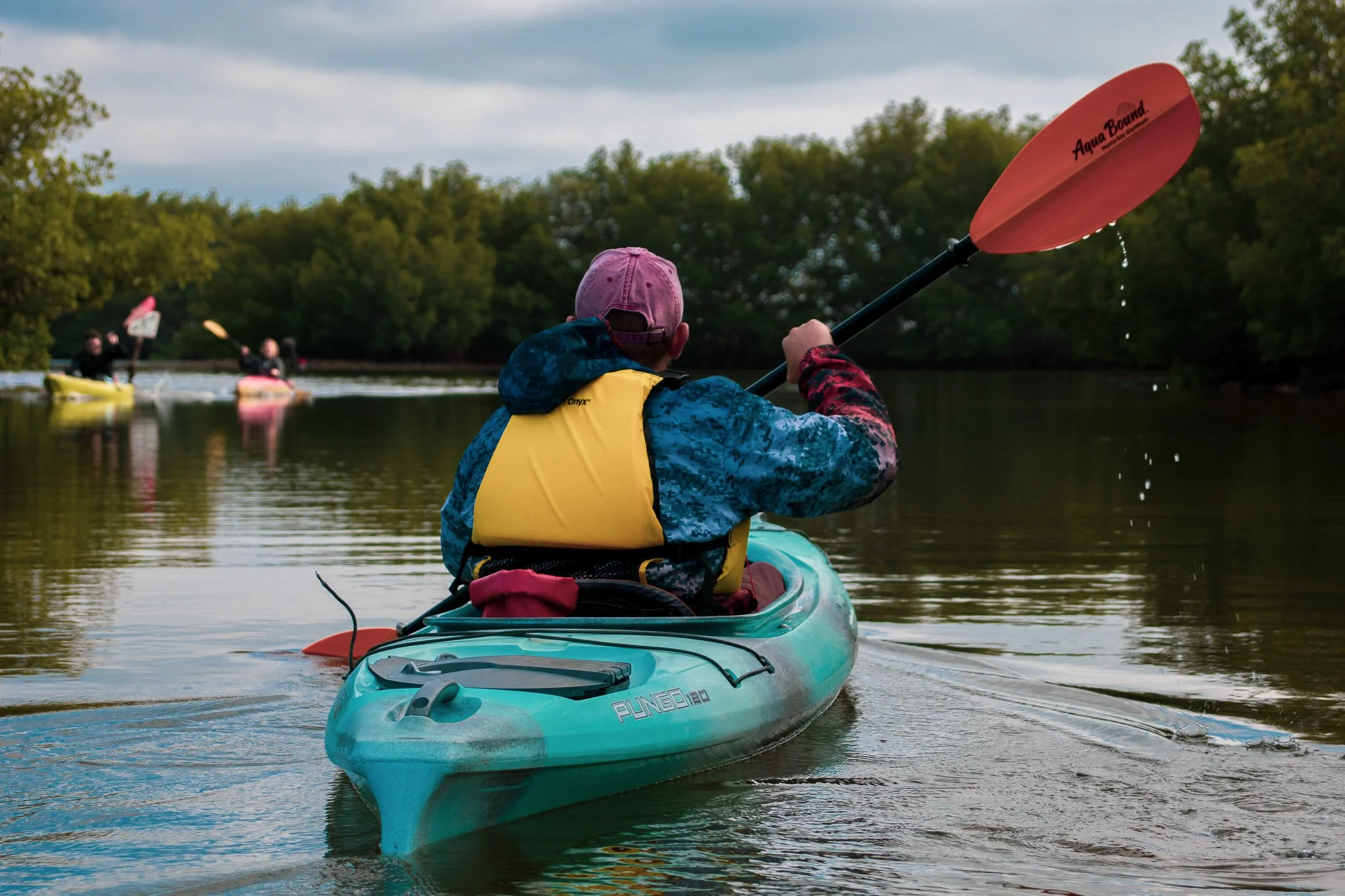 Person in a yellow life jacket and pink cap paddling a turquoise kayak on a calm waterway, with other kayakers and lush green trees in the background.