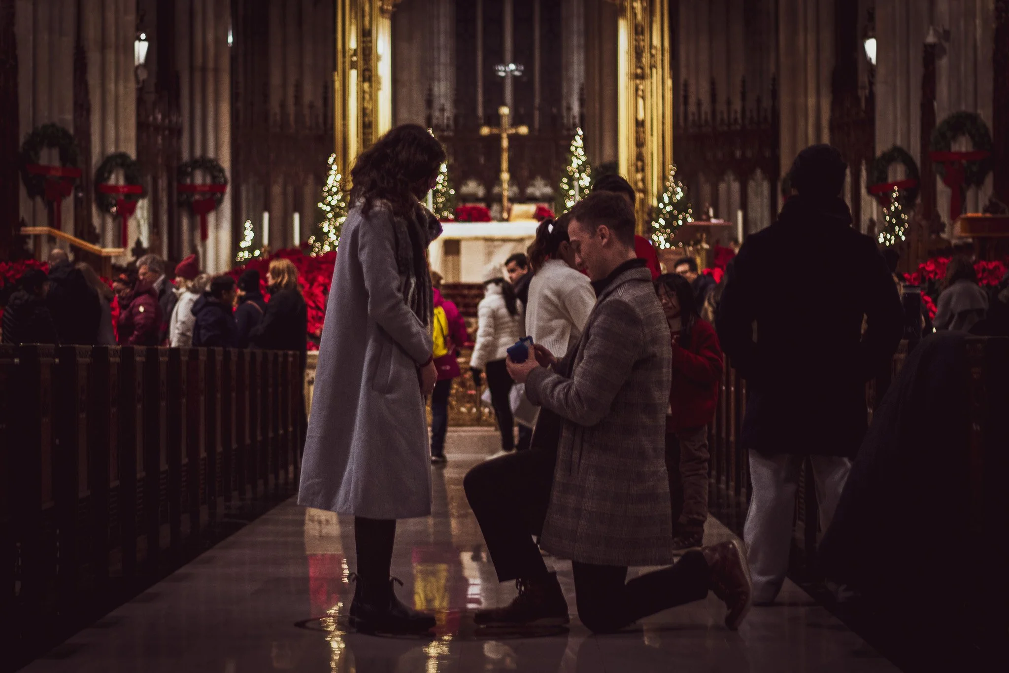 Kneeling man proposing to standing woman inside a church decorated for Christmas with poinsettias and Christmas trees.