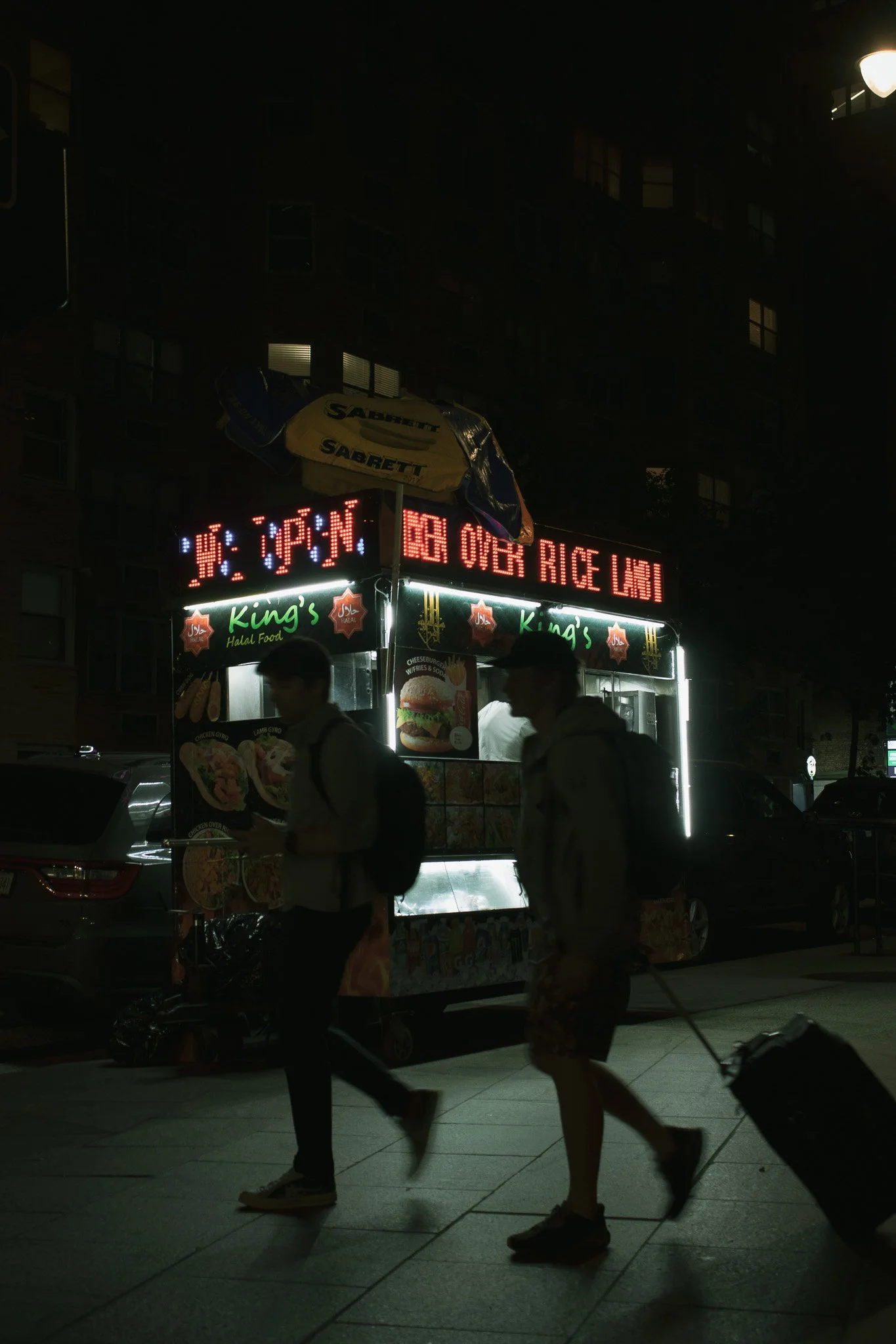 Night scene of a street food stall named 'King's' offering Halal food, with digital signs displaying menu items and prices. Pedestrians walk past, some carrying shopping bags or rolling luggage, with a residential building in the background.
